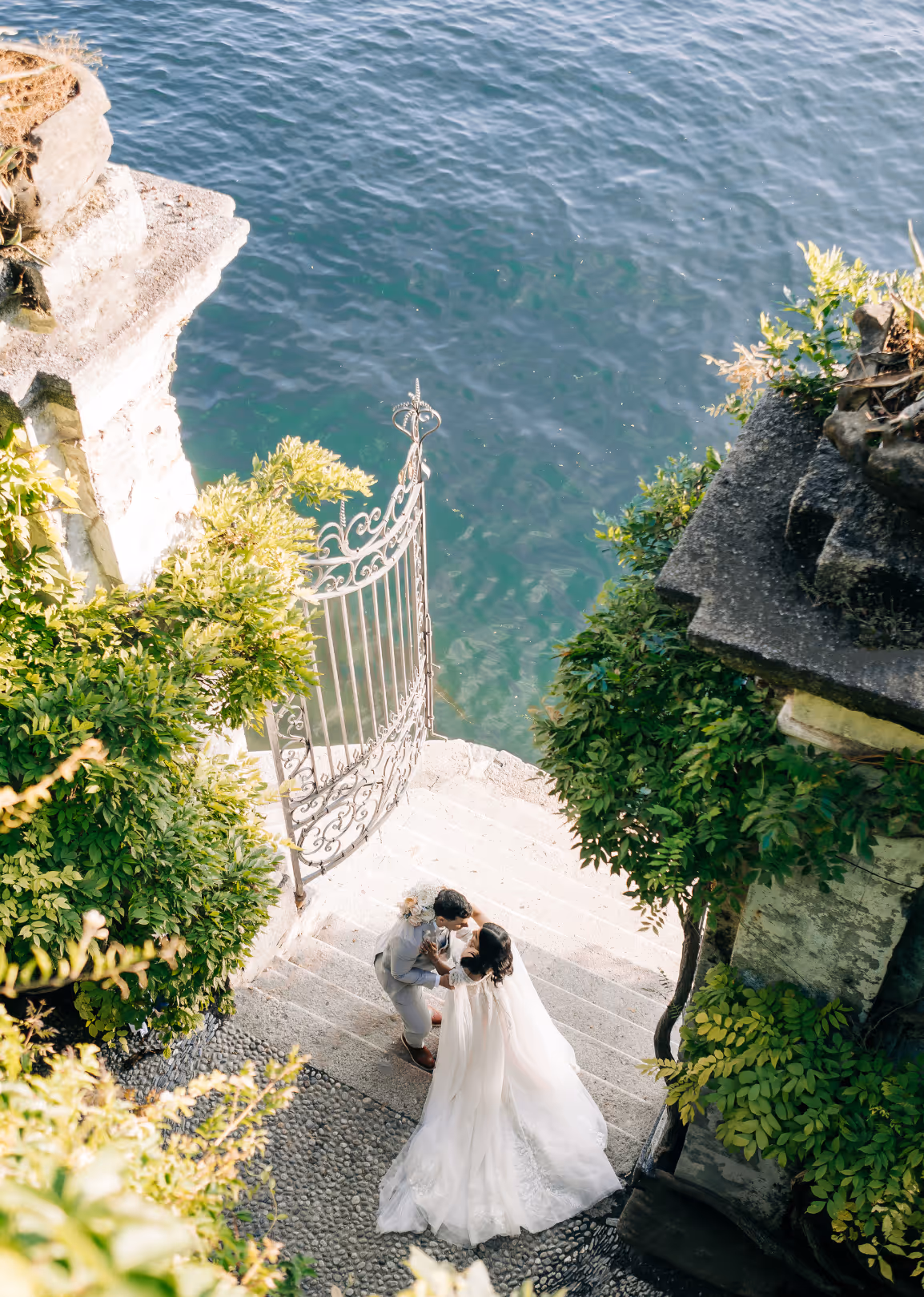 A high-angle vertical wedding photograph looking down at Puja and Rino embracing on a stone staircase next to an antique iron gate and the turquoise waters of Lake Como at Villa del Balbianello.