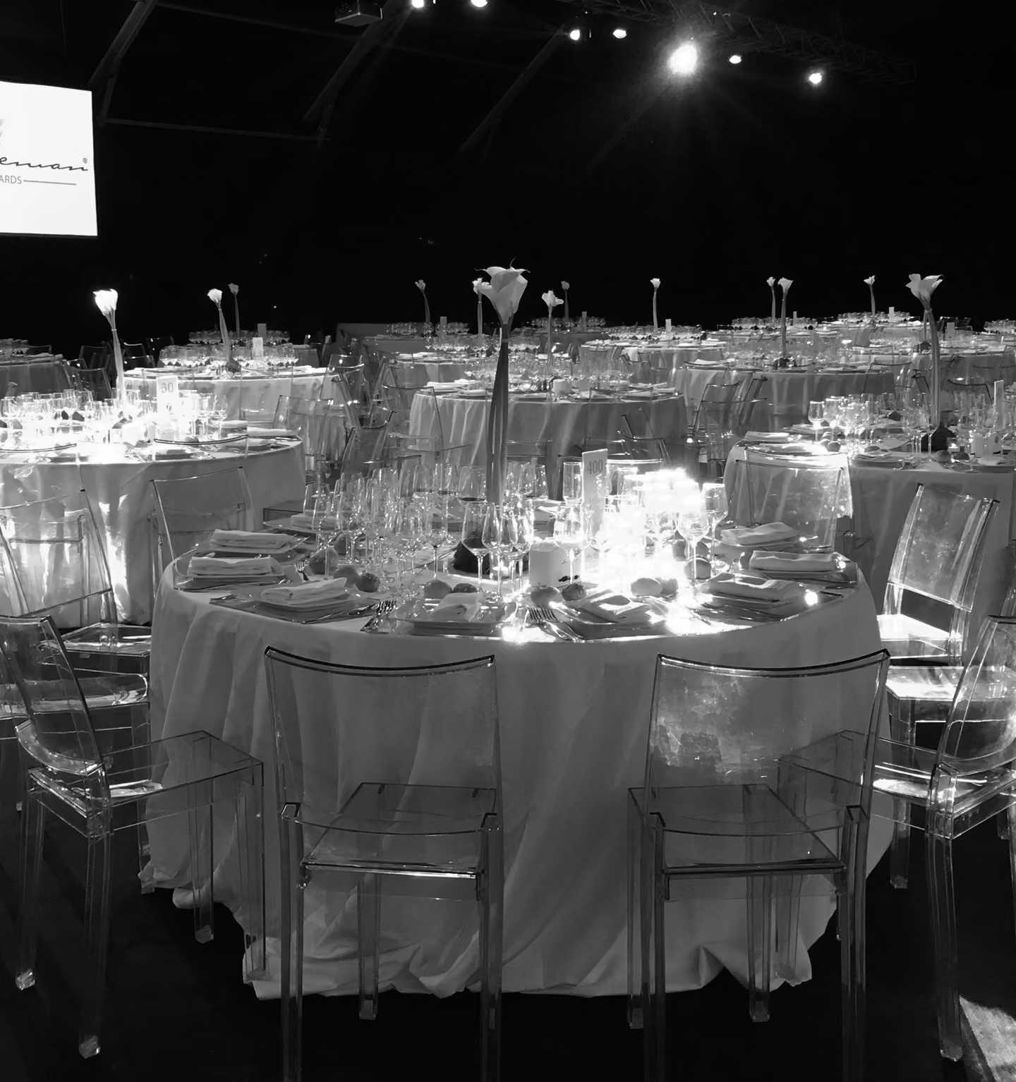 Sala allestita per la cena di gala del Premio Gentleman, caratterizzata da tavoli rotondi con tovaglie bianche, sedie trasparenti di design, calici di cristallo scintillanti e alti vasi minimalisti con calle bianche.