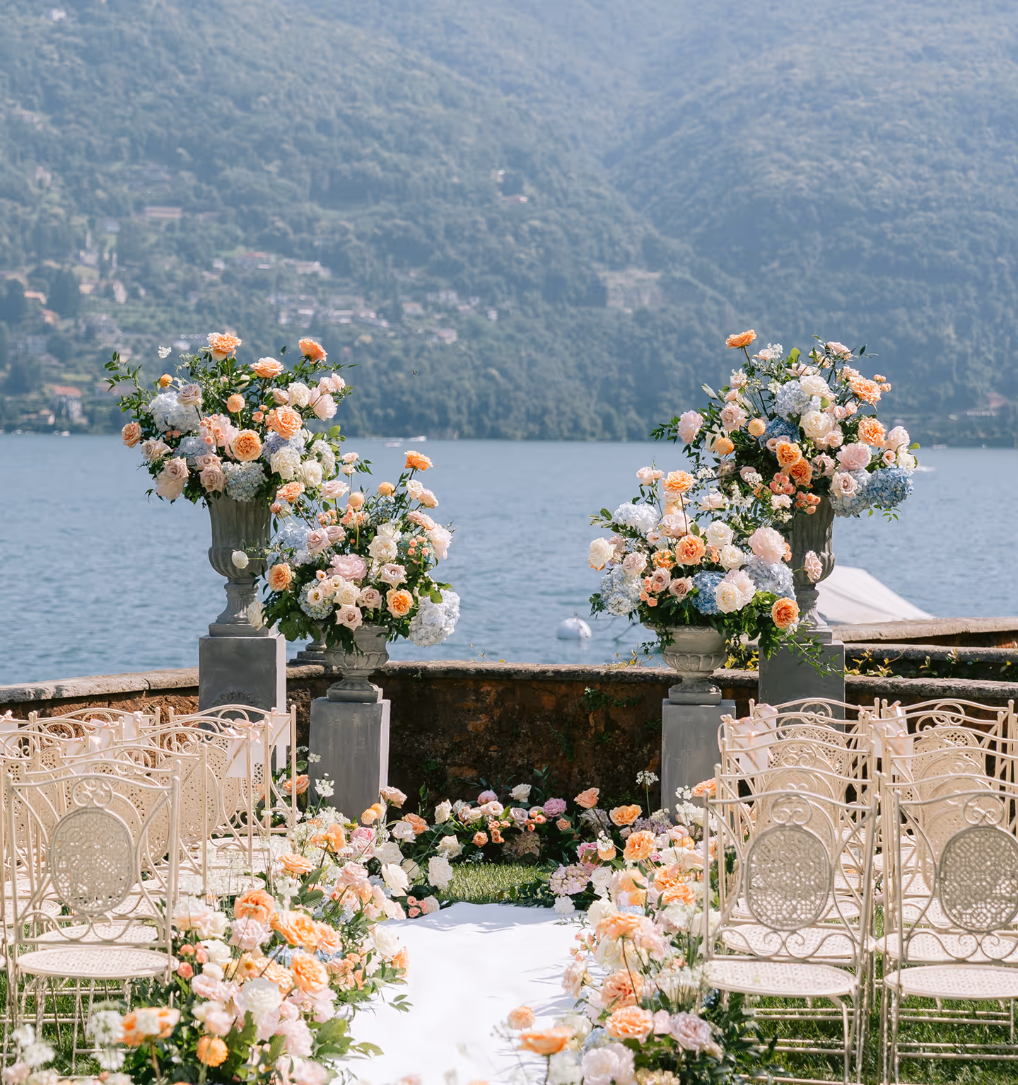 Navata di una cerimonia nuziale con sedie trasparenti e ricche composizioni di fiori color pastello in grandi vasi di pietra, allestita direttamente sul lungolago di Villa Regina Teodolinda con le montagne di Como sullo sfondo.