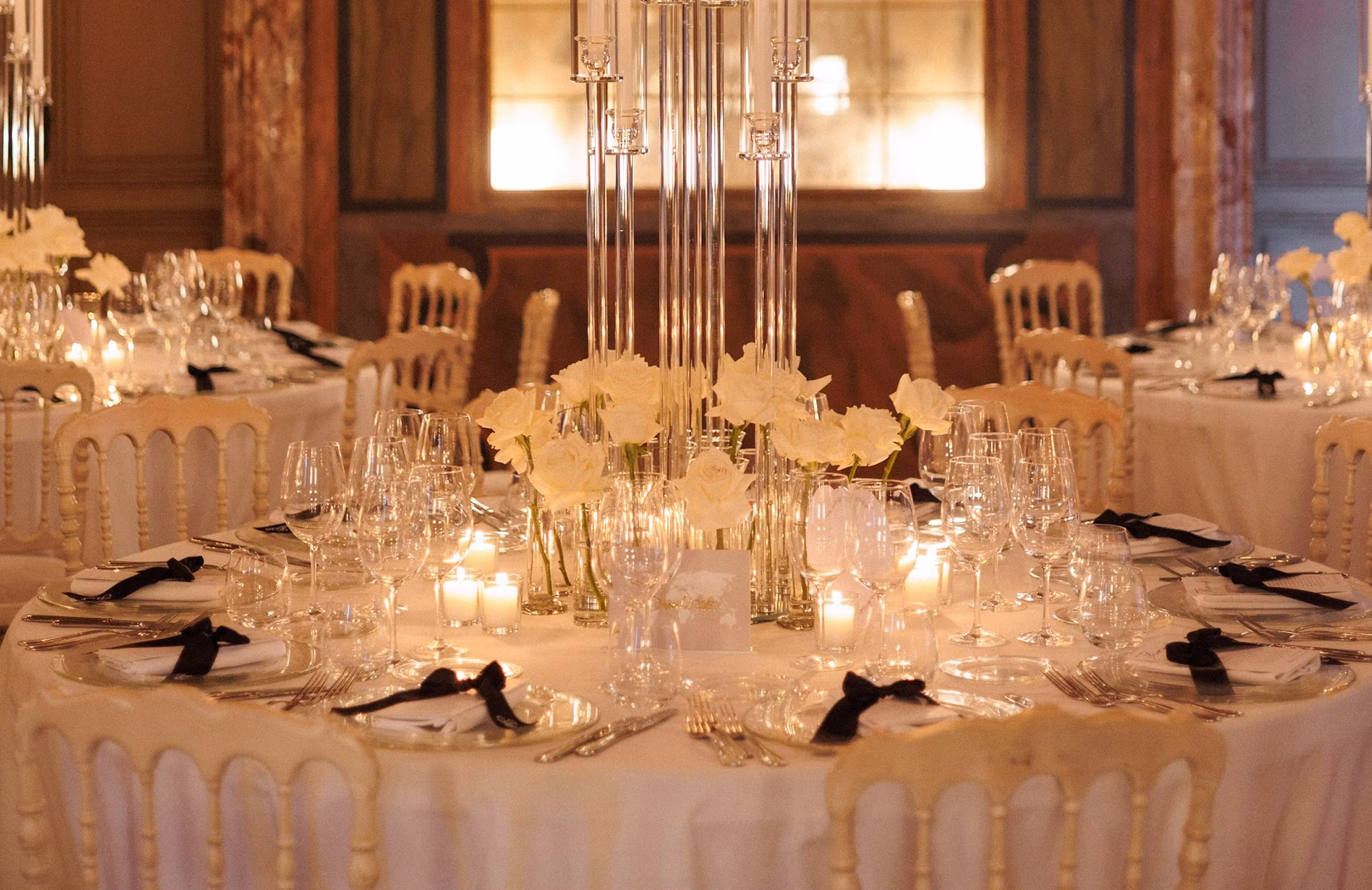 Dettaglio di un tavolo tondo elegantemente apparecchiato per il ricevimento di matrimonio nei saloni di Villa Erba, con un grande candelabro di cristallo trasparente, rose bianche, candele accese e tovaglioli decorati con un fiocco nero.