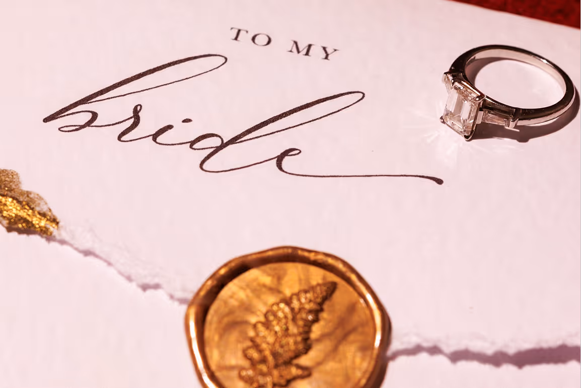 Dettaglio ravvicinato di un prezioso anello di fidanzamento con diamante solitario adagiato su una lettera d'amore artigianale con la scritta calligrafica "To my bride" e un sigillo dorato.