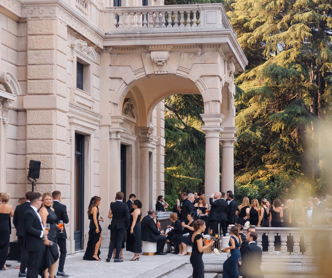 Un gruppo di ospiti in abito da sera elegante nero partecipa a un cocktail party all'aperto sulla terrazza in pietra di una storica villa monumentale con porticato ad archi.