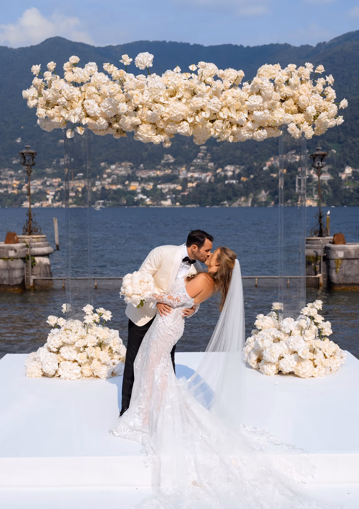 Lo sposo in smoking bianco bacia la sposa stringendola in un romantico casqué sull'altare fronte lago di Villa Erba, sotto una spettacolare composizione sospesa di rose e fiori bianchi.