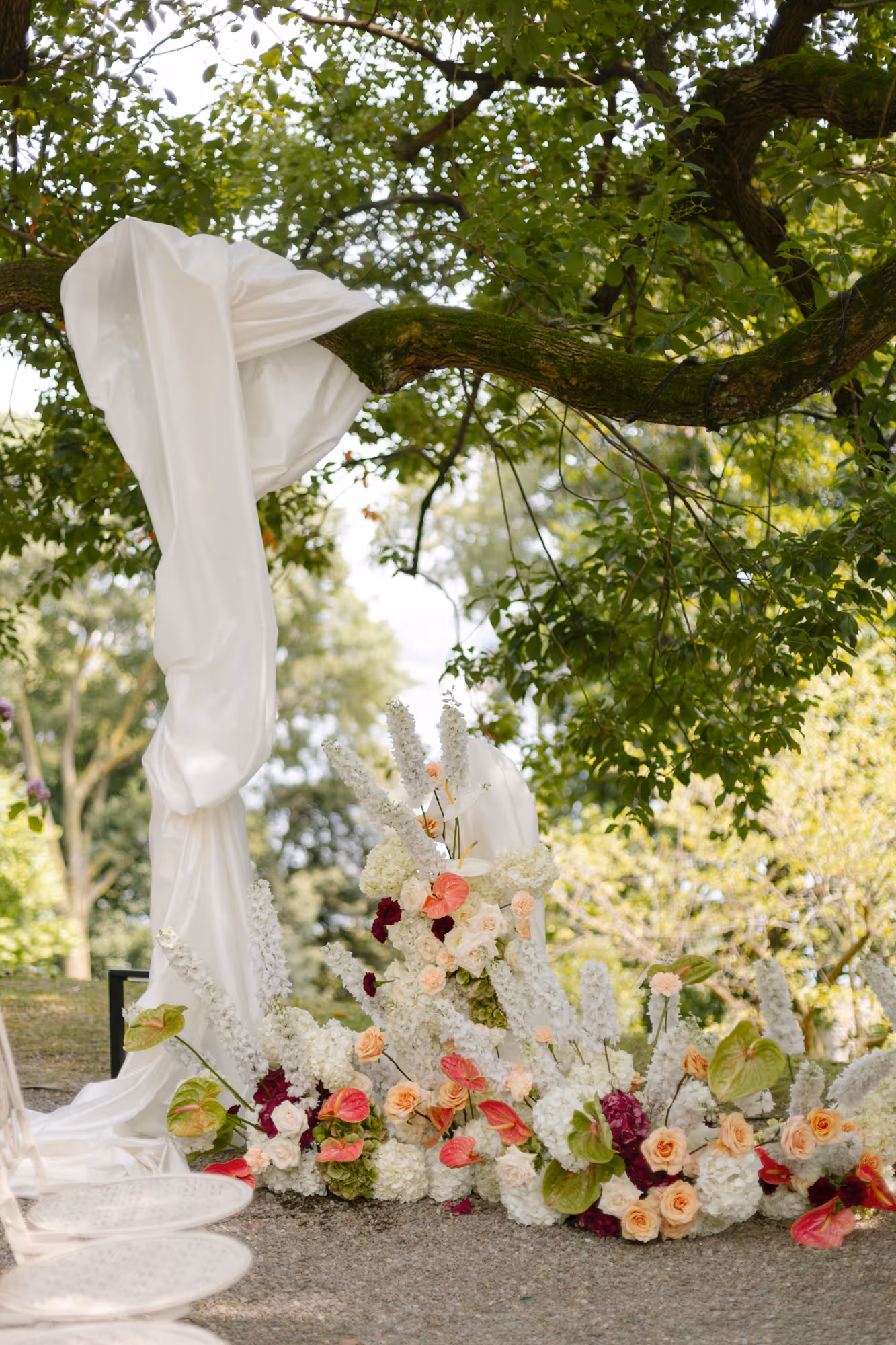 A botanical outdoor wedding altar at Villa Grumello featuring elegant white fabric draped over a mossy tree branch and lush ground floral arrangements with roses, hydrangeas, and pink anthuriums.
