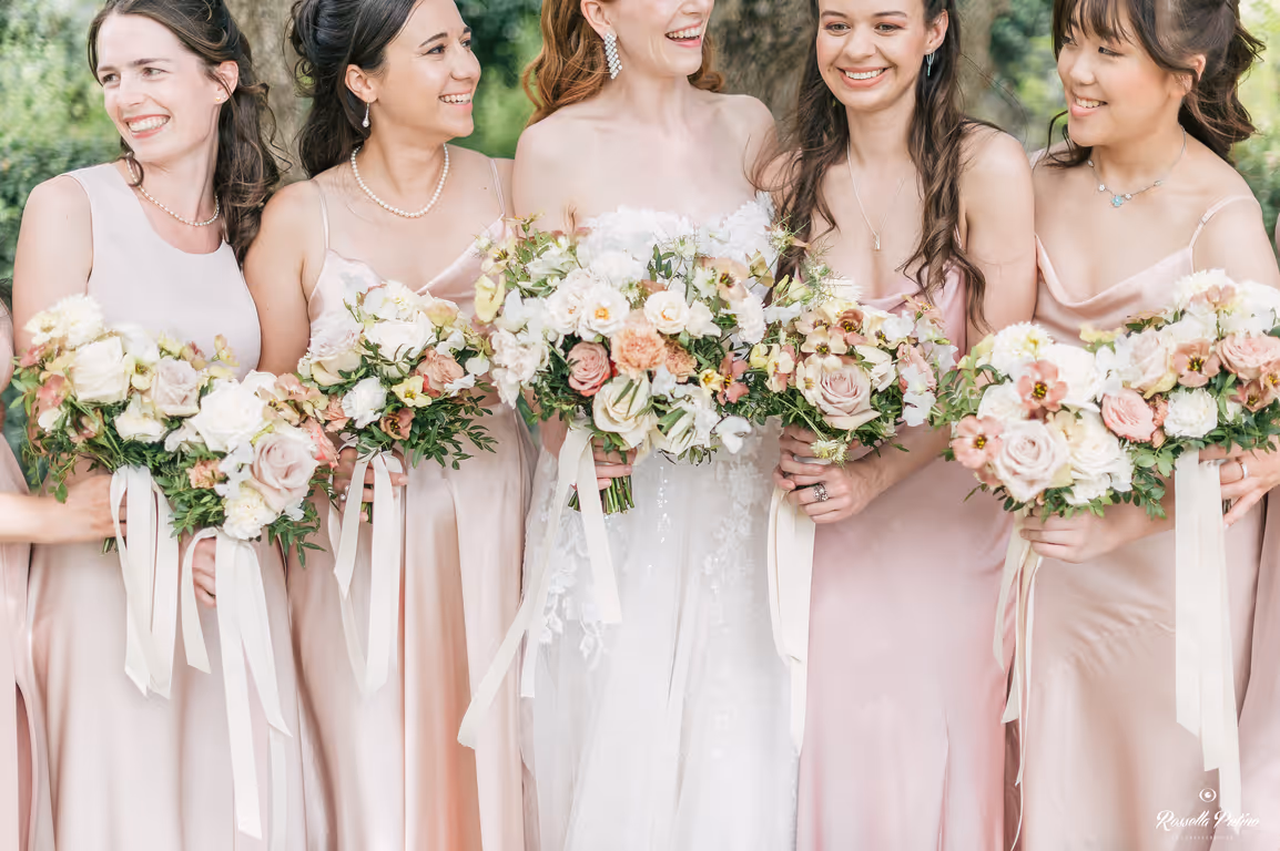 A close-up crop of a bride and her bridesmaids in elegant silk blush pink gowns, holding matching fine-art wedding bouquets with pastel roses and long ribbons at Castello di Vicarello.