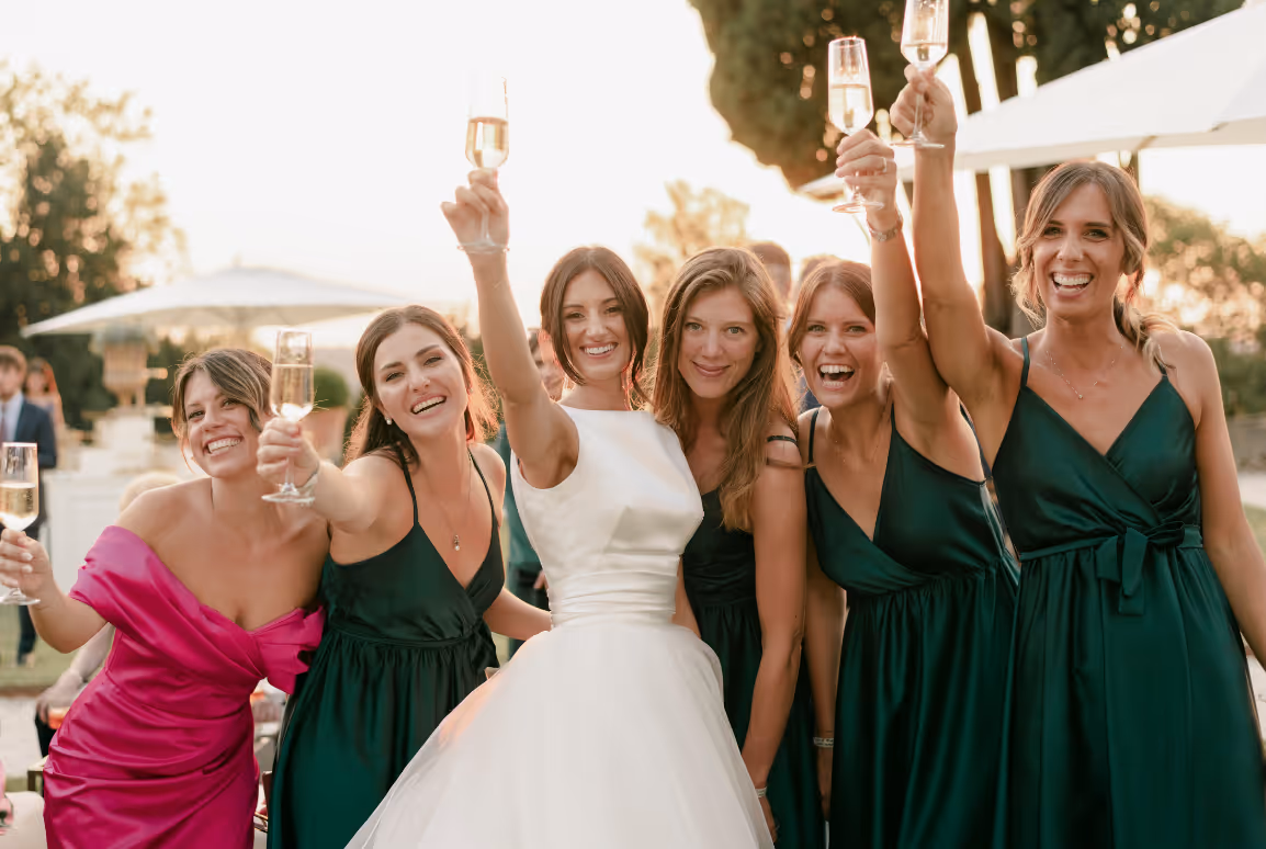 A joyful horizontal photograph of the bride, Martina, cheering and raising champagne glasses with her bridesmaids dressed in emerald green at Castello Durini.