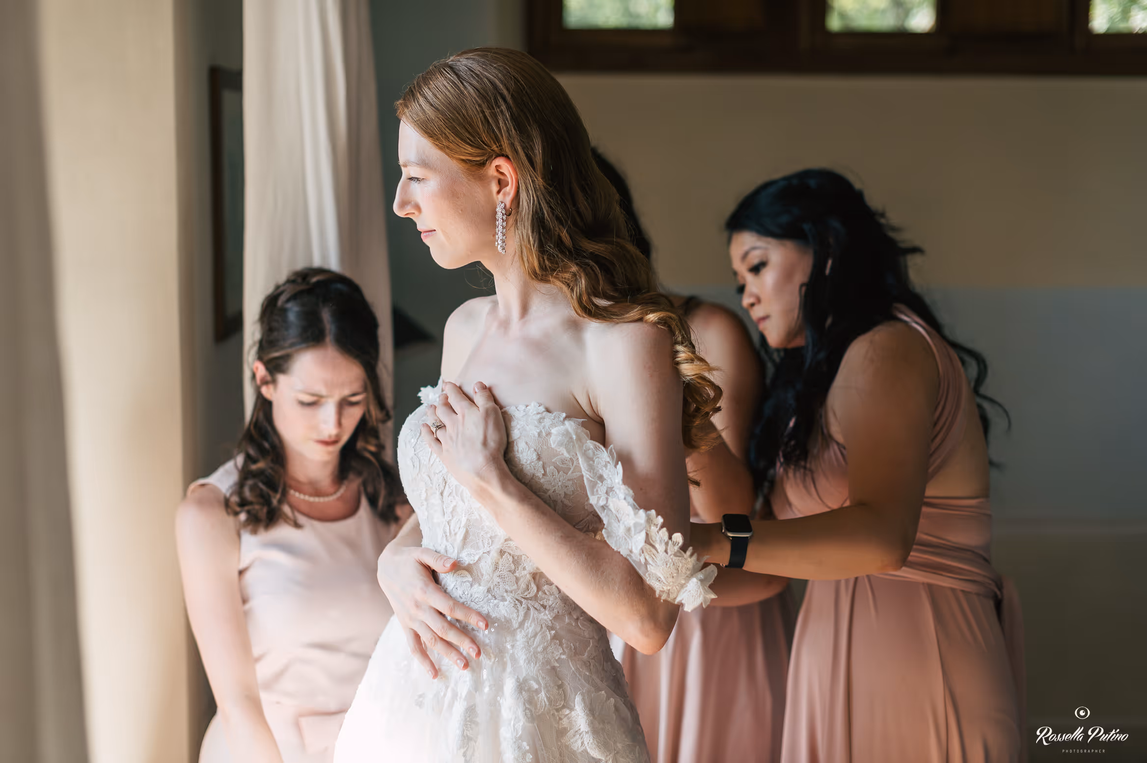 An intimate indoor photograph of a bride in a strapless lace wedding gown getting ready with the help of her bridesmaids in dusty pink dresses at Castello di Vicarello.