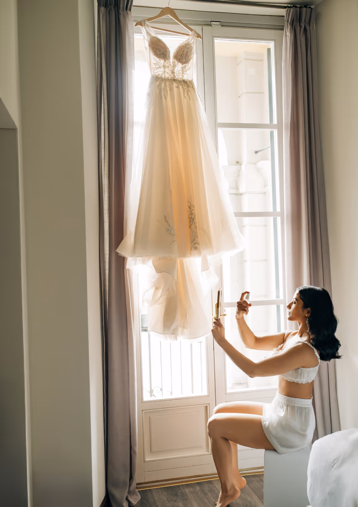 A vertical indoor photograph of the bride, looking up at her detailed lace wedding gown hanging elegantly against a large sunlit window during preparations at Villa Balbianello.