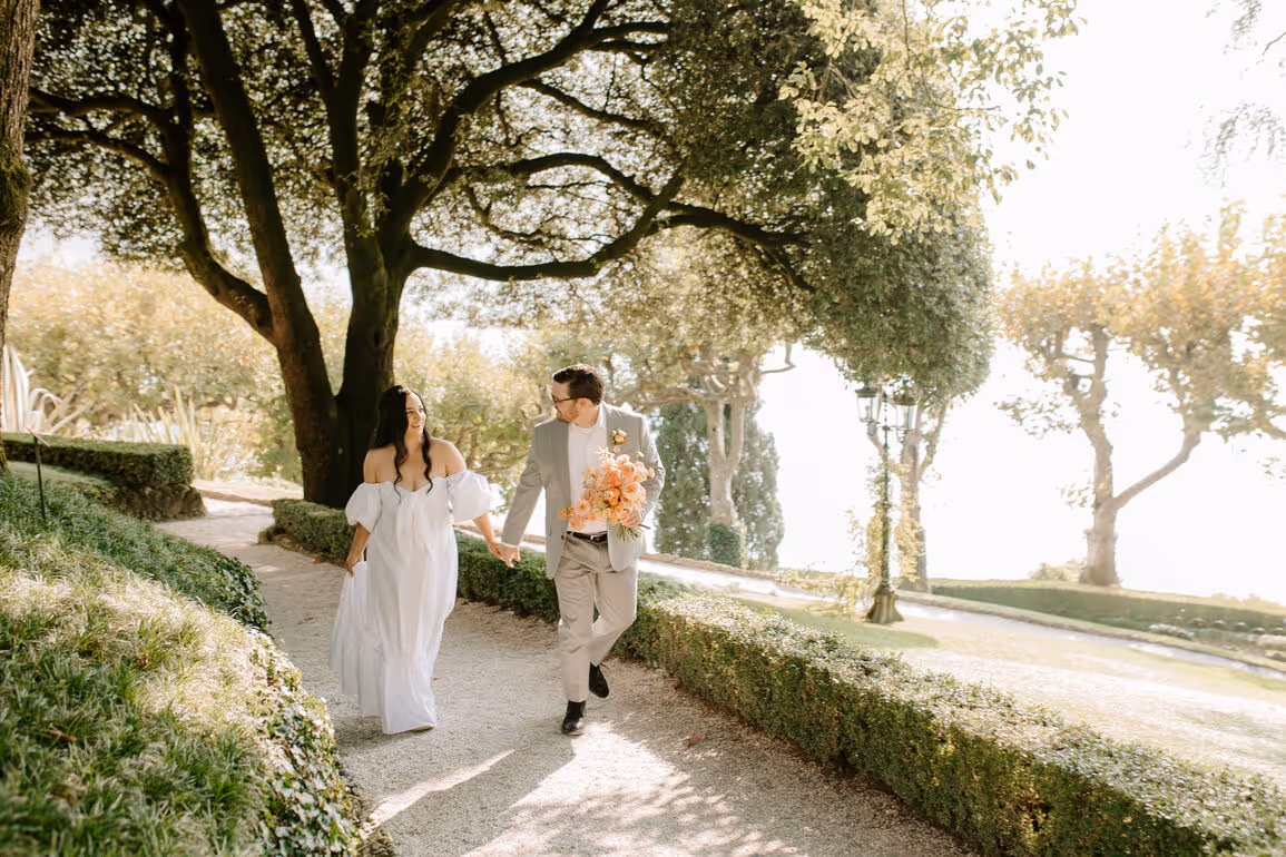 A bright landscape photograph of a bride and groom holding hands while walking along a manicured garden pathway under large trees at Villa del Balbianello.