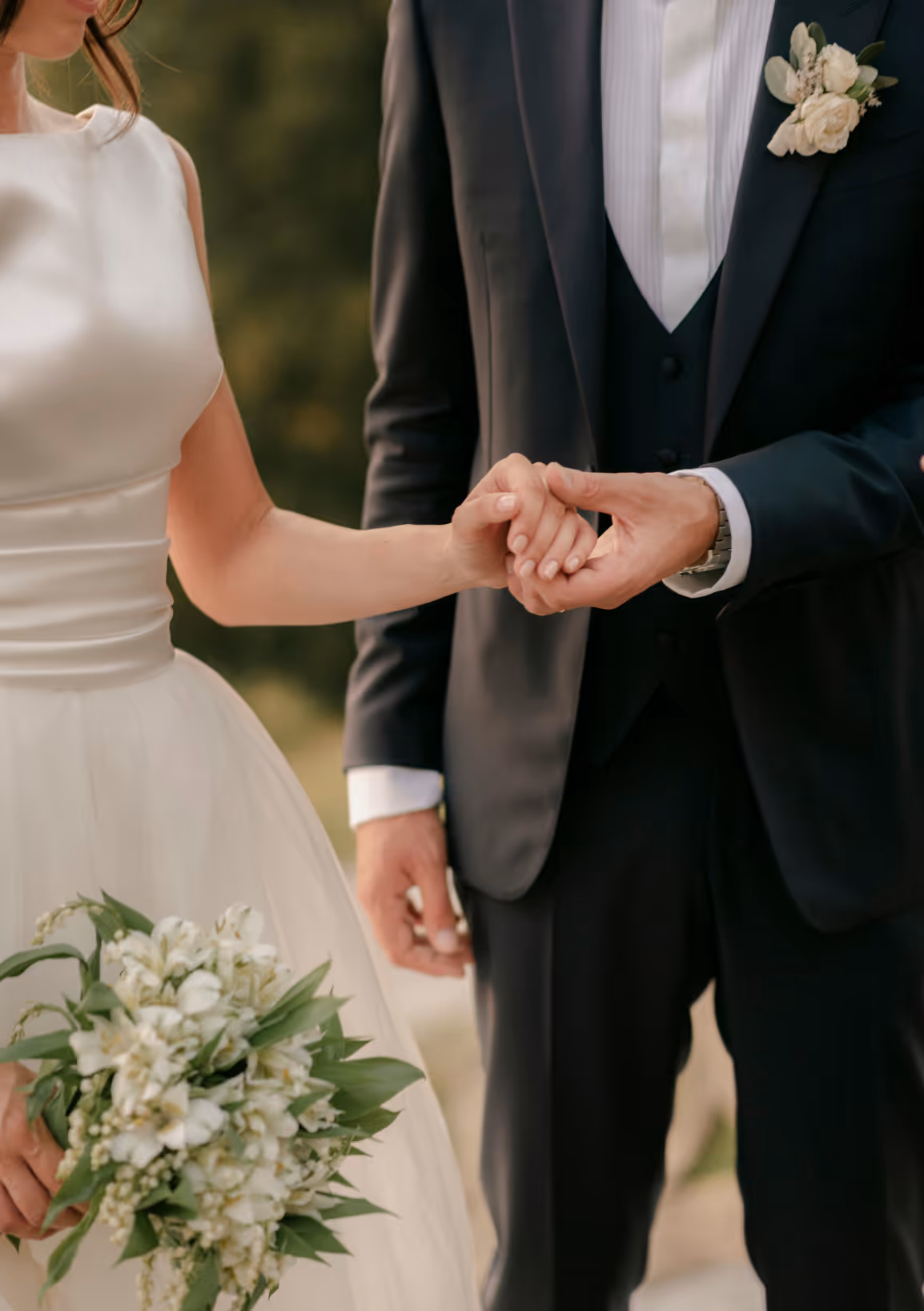 A vertical detail photograph of a groom in a dark blue suit holding his bride's hand, showcasing her elegant satin wedding dress and his white rose boutonniere.