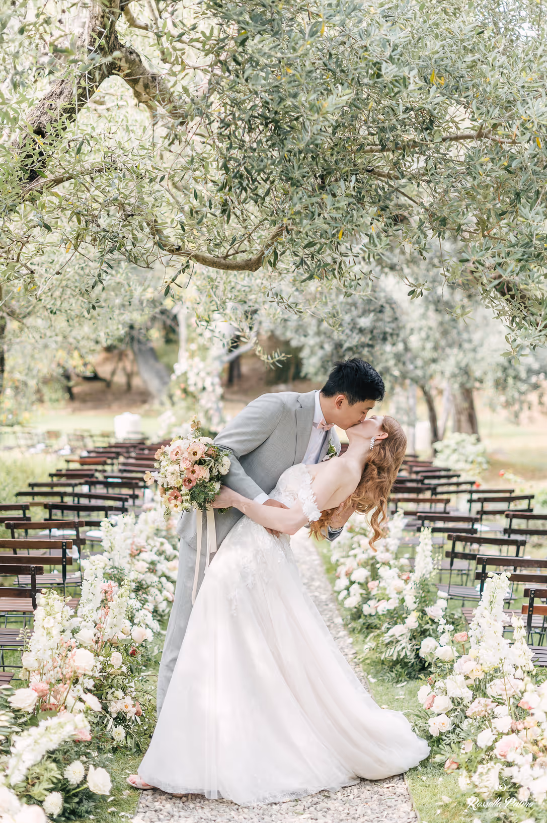 A vertical portrait of a groom dipping and kissing his bride along a gravel wedding aisle lined with white roses and delphiniums under the shade of olive trees at Castello di Vicarello.