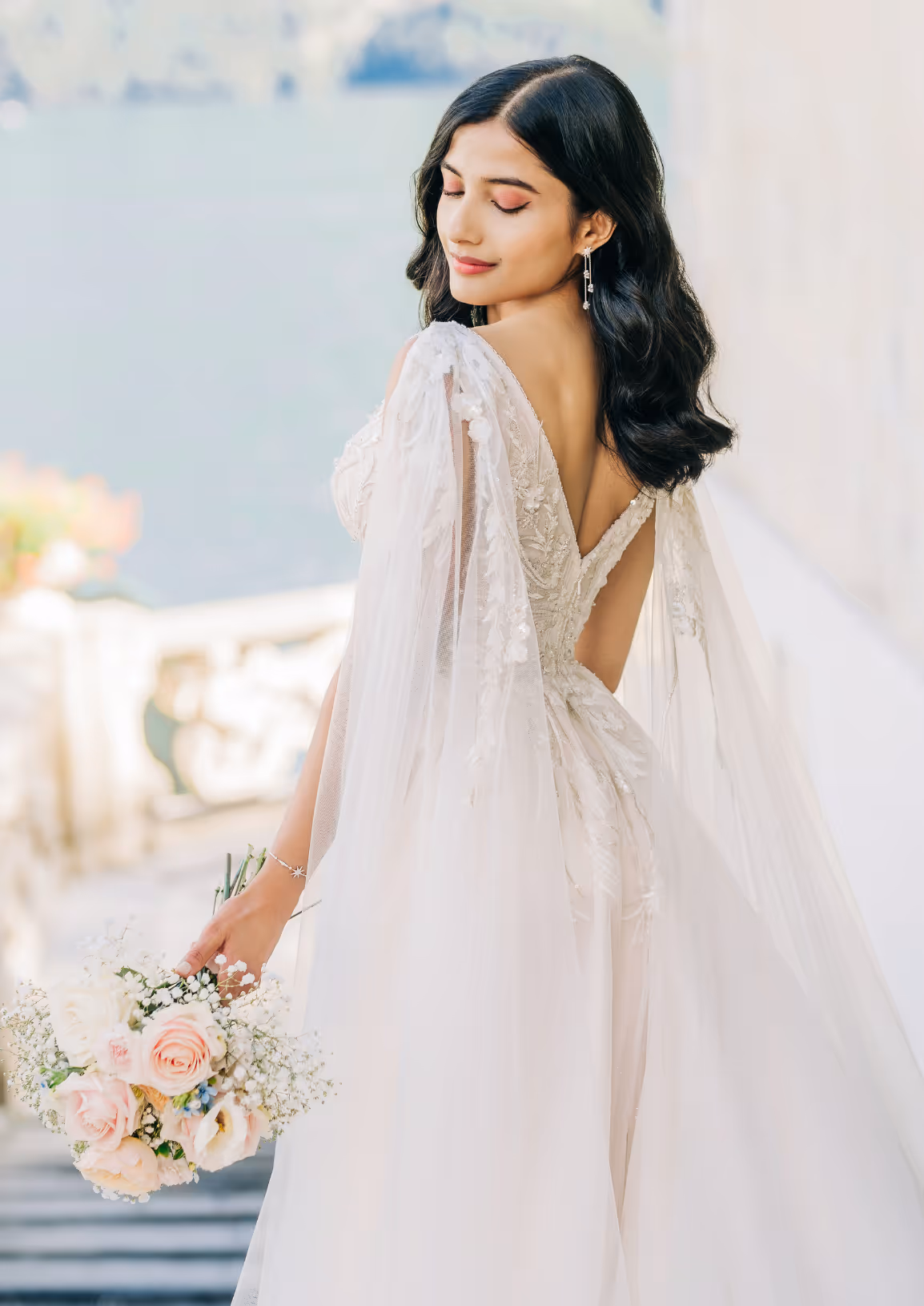 A vertical fine art bridal portrait of Puja showing the detailed V-back and tulle shoulder capes of her wedding gown while holding a blush pink bouquet at Villa del Balbianello.
