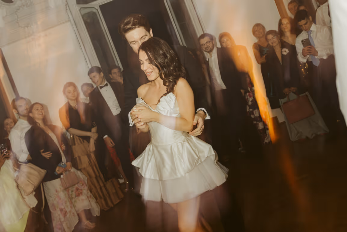 A colorful, warm-toned reception photo with an artistic light leak effect, showing the bride in a playful short satin party dress smiling next to her husband while guests watch the celebration.