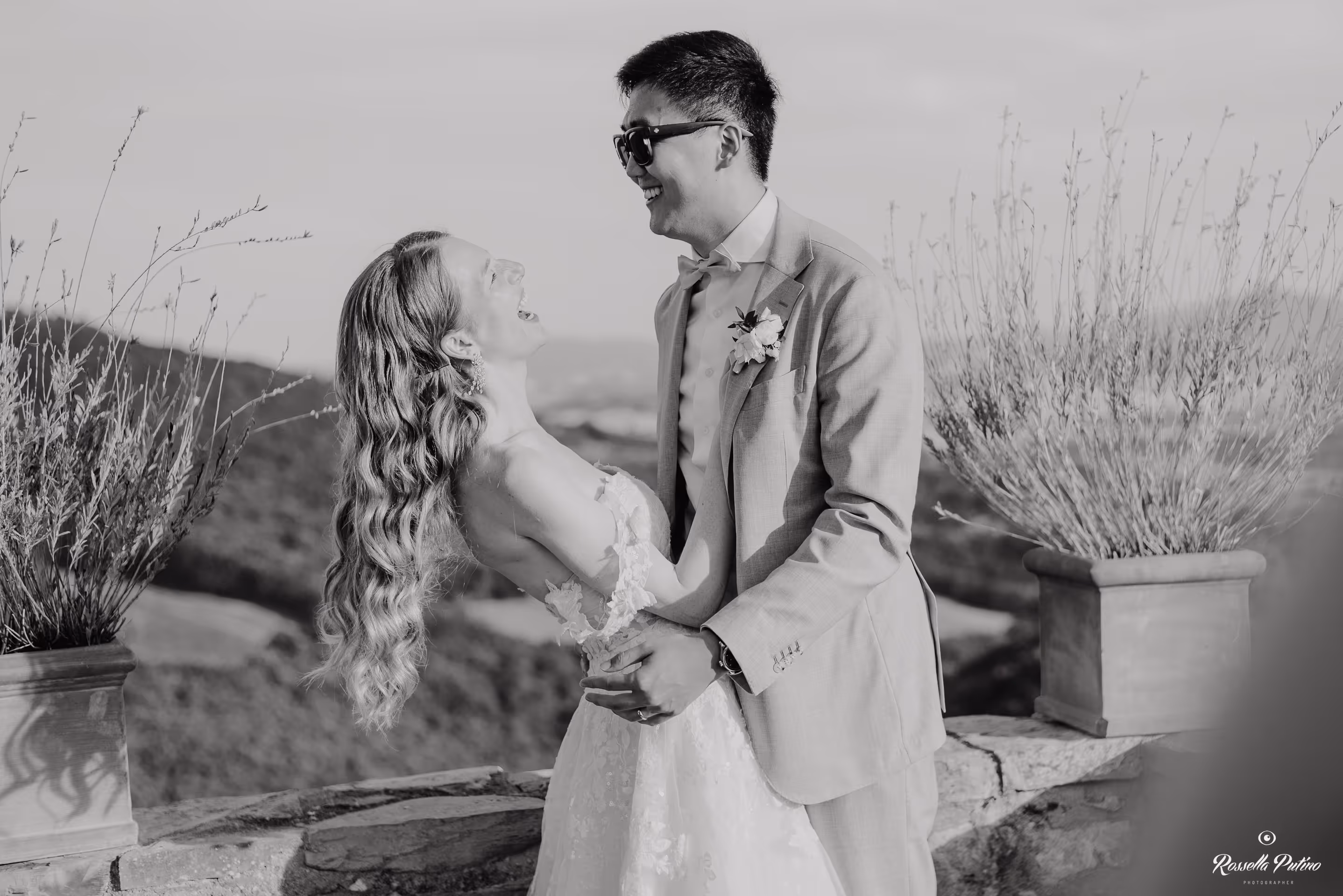 A joyful black and white candid portrait of a bride laughing with her head tilted back while embracing her husband near a stone wall at Castello di Vicarello.