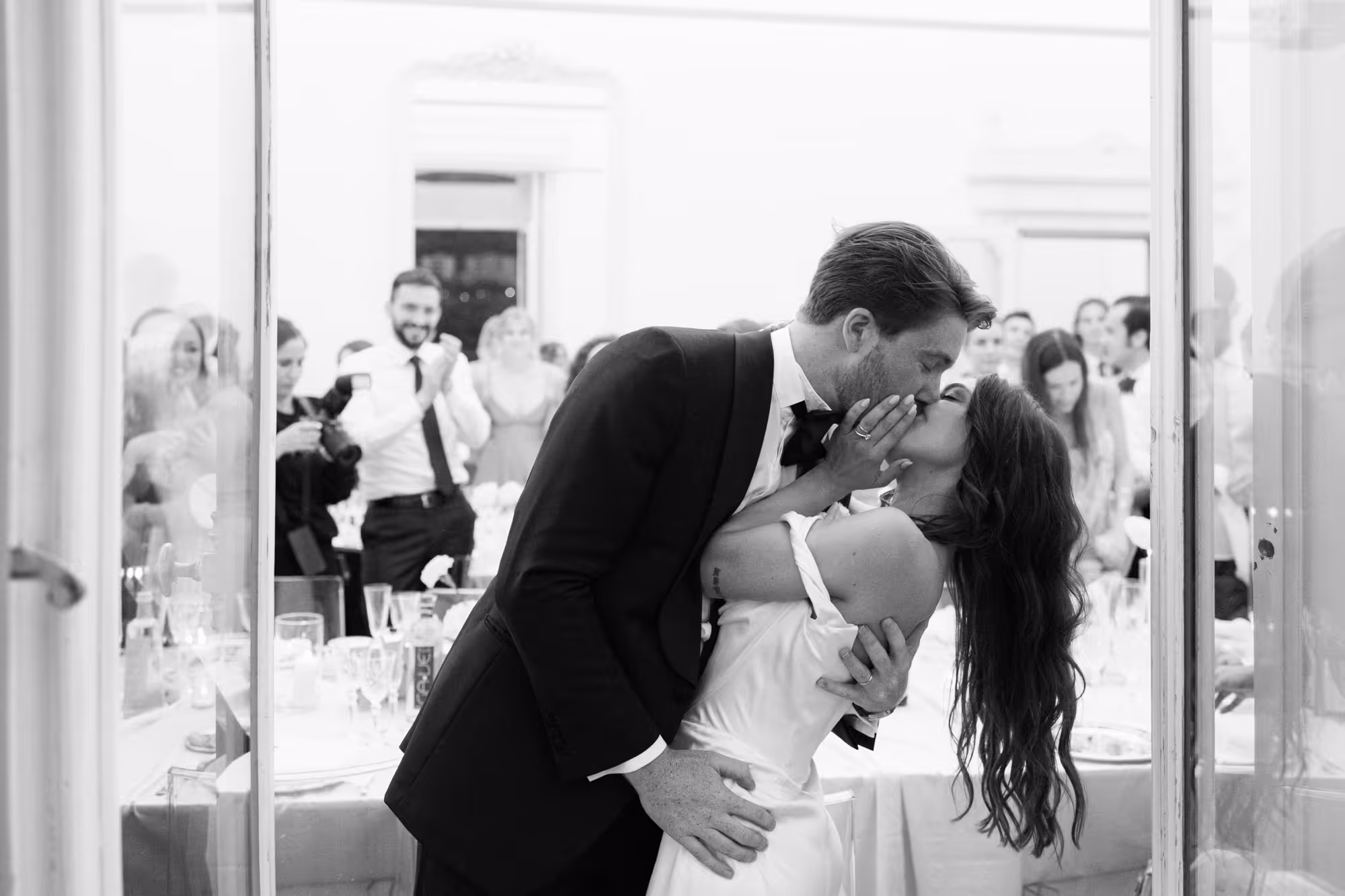 A candid black and white photo of a groom dipping his bride for a romantic kiss during their wedding reception dinner inside Villa Grumello, with guests applauding in the background.