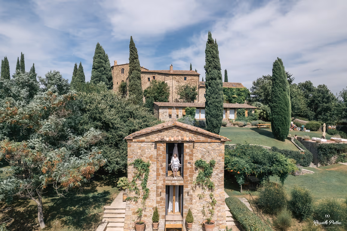 A wide landscape photograph of the stone buildings and rustic outhouses at Castello di Vicarello, framed by iconic green cypress trees under a blue, cloudy sky.