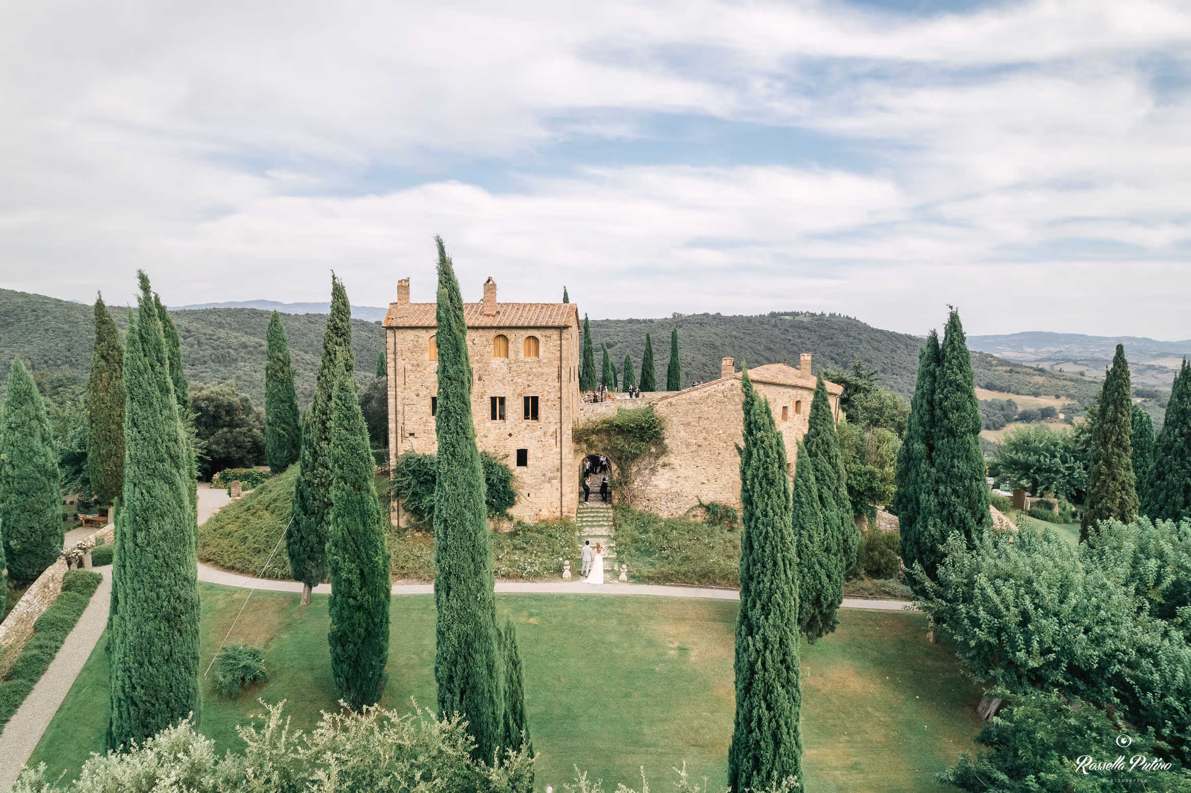 A wide scenic view of Castello di Vicarello in Tuscany during a destination wedding, featuring the historic stone castle surrounded by iconic tall cypress trees and rolling green hills.
