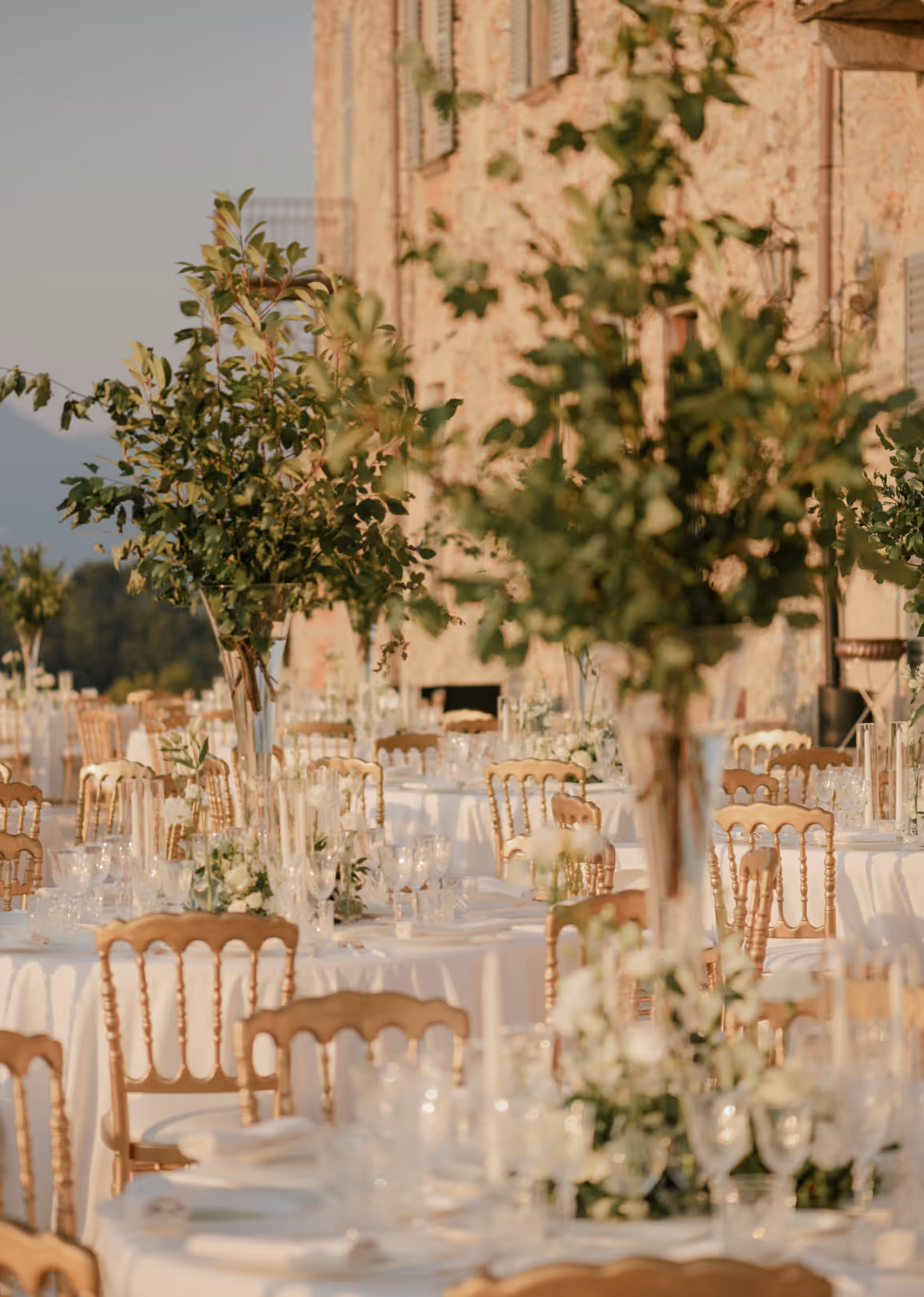 A vertical detail shot of an outdoor wedding reception at Castello Durini featuring tall glass vases with leafy green branches on tables with gold chairs along the stone villa facade.