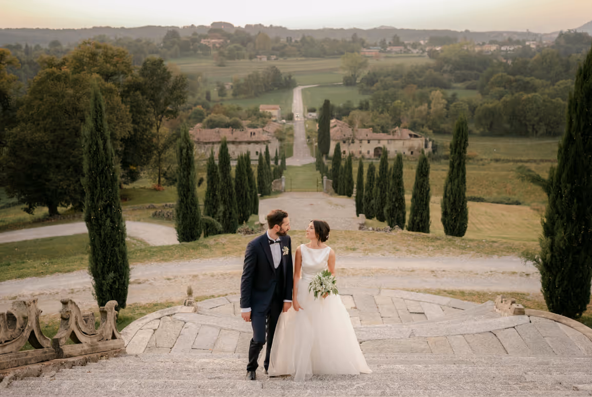 A panoramic wedding cover photo of bride and groom about to kiss, looking over a valley framed by a grand avenue of cypress trees at Castello Durini in Italy.