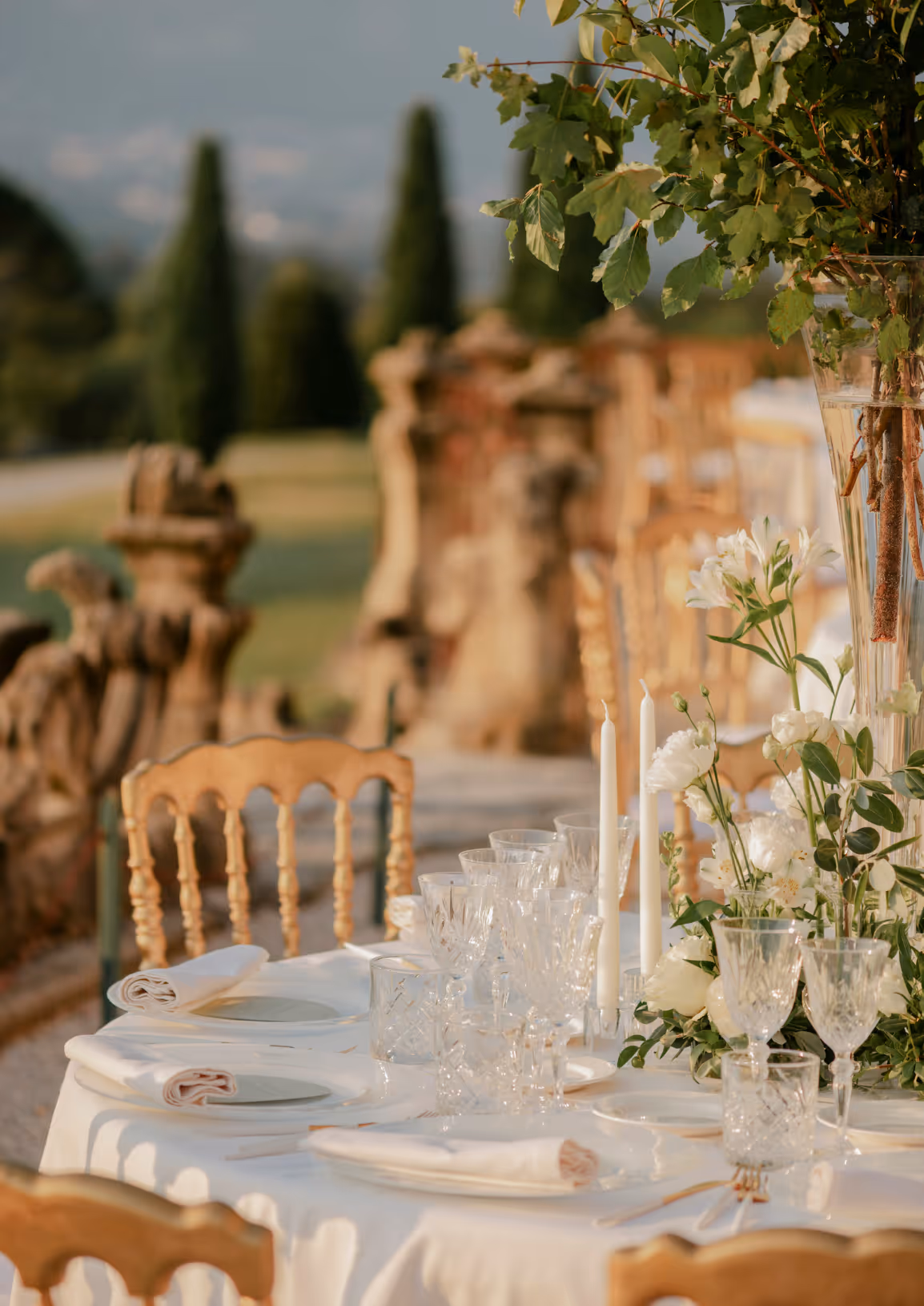 A close-up vertical photograph of a wedding reception dining table at Castello Durini featuring elegant crystal glassware, white taper candles, and white flowers in the golden afternoon sun.
