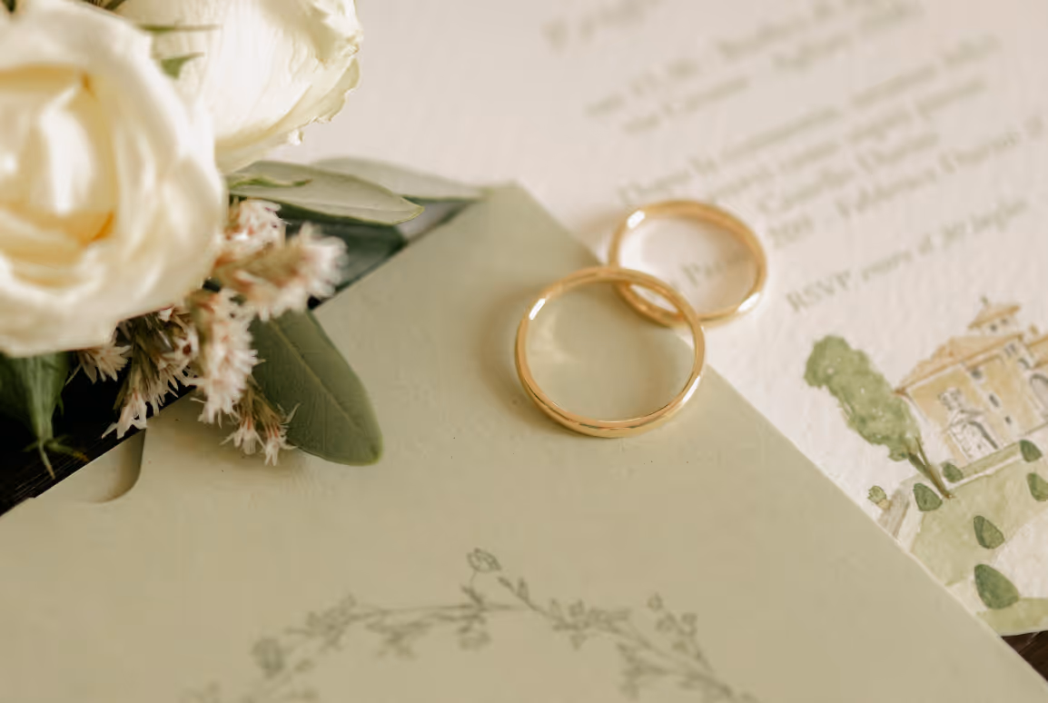 A close-up horizontal photograph of gold wedding bands resting on an illustrated fine art wedding invitation next to an olive green envelope and white roses.