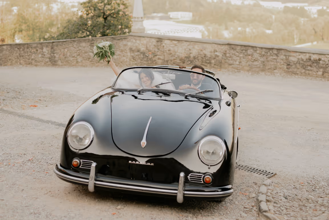 A landscape photograph of a bride and groom driving away in a classic black Porsche speedster convertible, with the bride cheering and lifting her bouquet at Castello Durini.