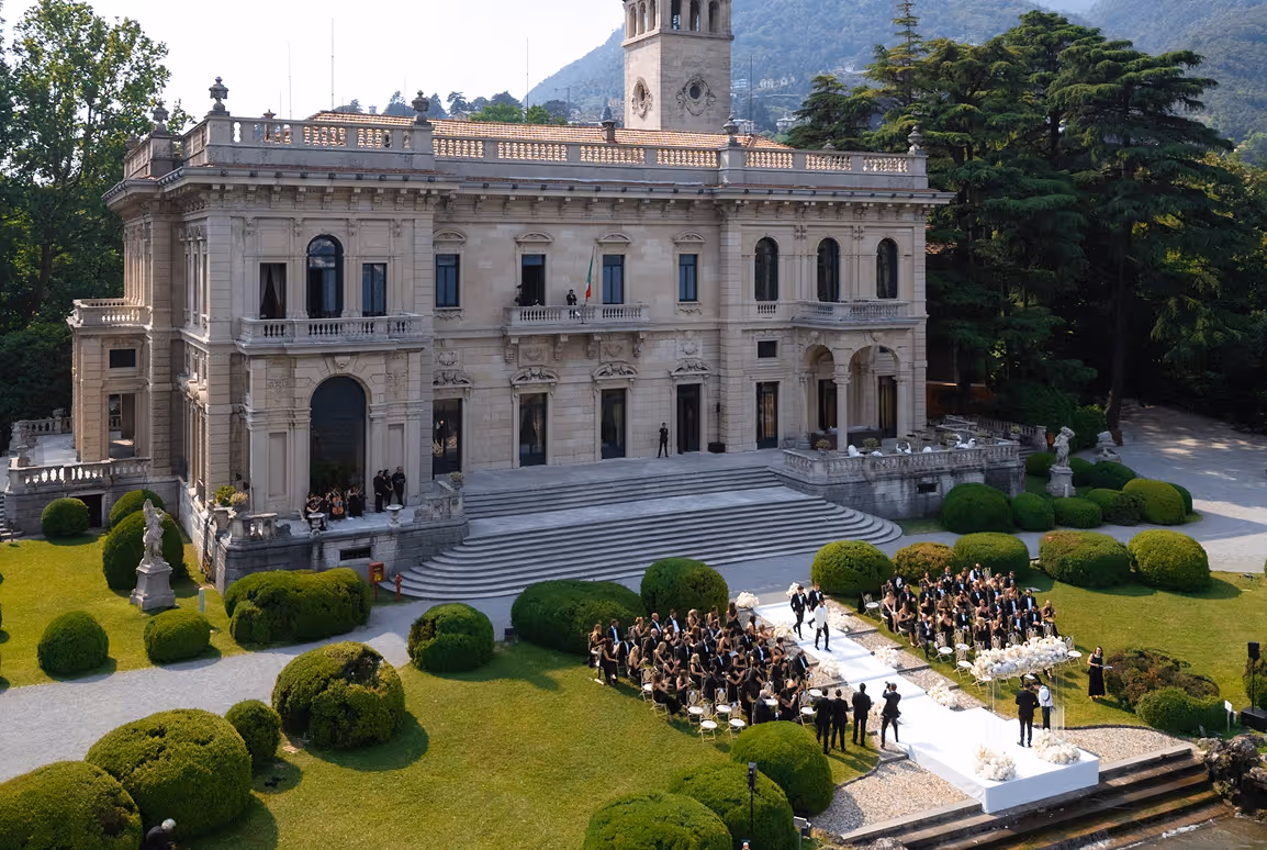 Veduta aerea dall'alto della cerimonia nuziale nel parco di Villa Erba, con gli ospiti seduti ai lati di una lunga passerella bianca che conduce all'altare fronte lago.