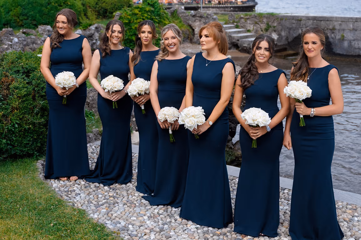 Sette damigelle d'onore in eleganti abiti lunghi coordinati color blu notte, sorridenti con in mano bouquet di rose bianche sul lungolago in sasso di Villa Erba.