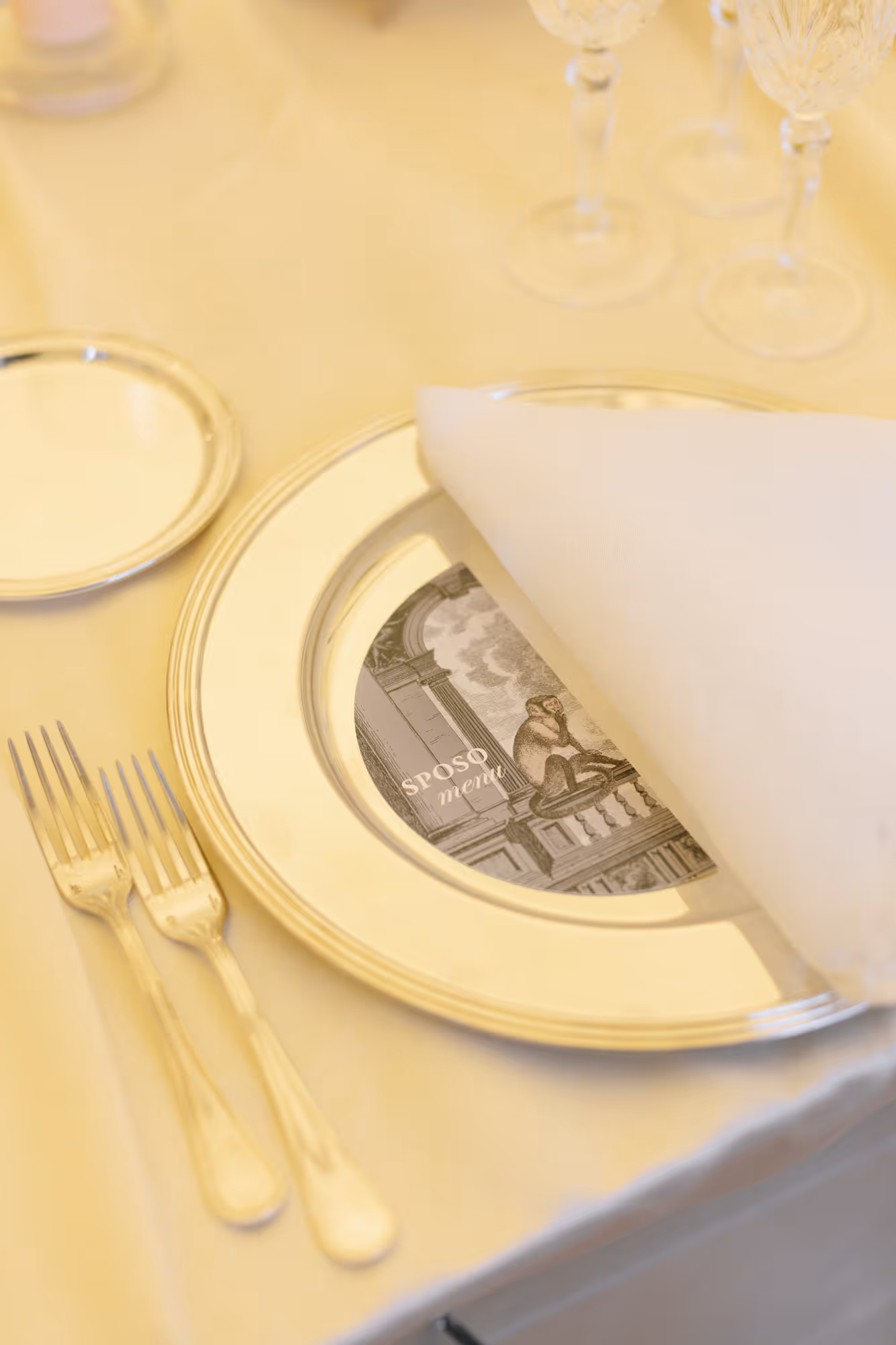 A close-up vertical shot of an elegant wedding table setting featuring a shiny silver charger plate, a custom illustrated menu card hidden under a white folded napkin, crystal glasses, and silver forks.