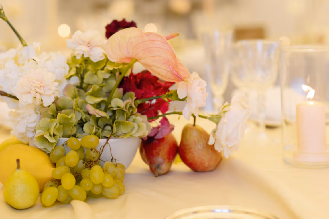 A beautiful fine art wedding centerpiece combining fresh green grapes, pears, a lemon, and delicate floral arrangements on a soft white tablecloth with a candle glowing in the background.