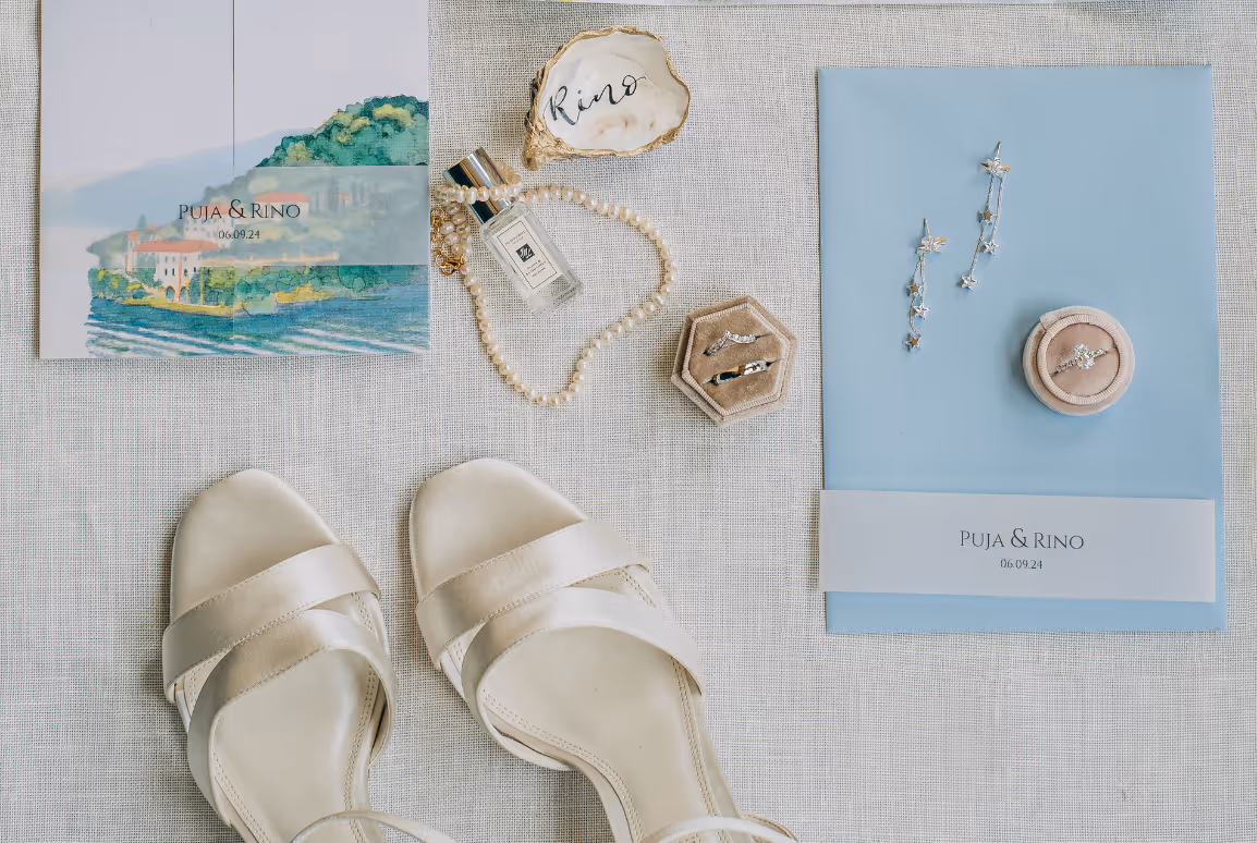 An overhead flat lay of destination wedding details for Puja and Rino, including an illustrated Lake Como invitation suite, white bridal shoes, jewelry, and a custom painted oyster shell.