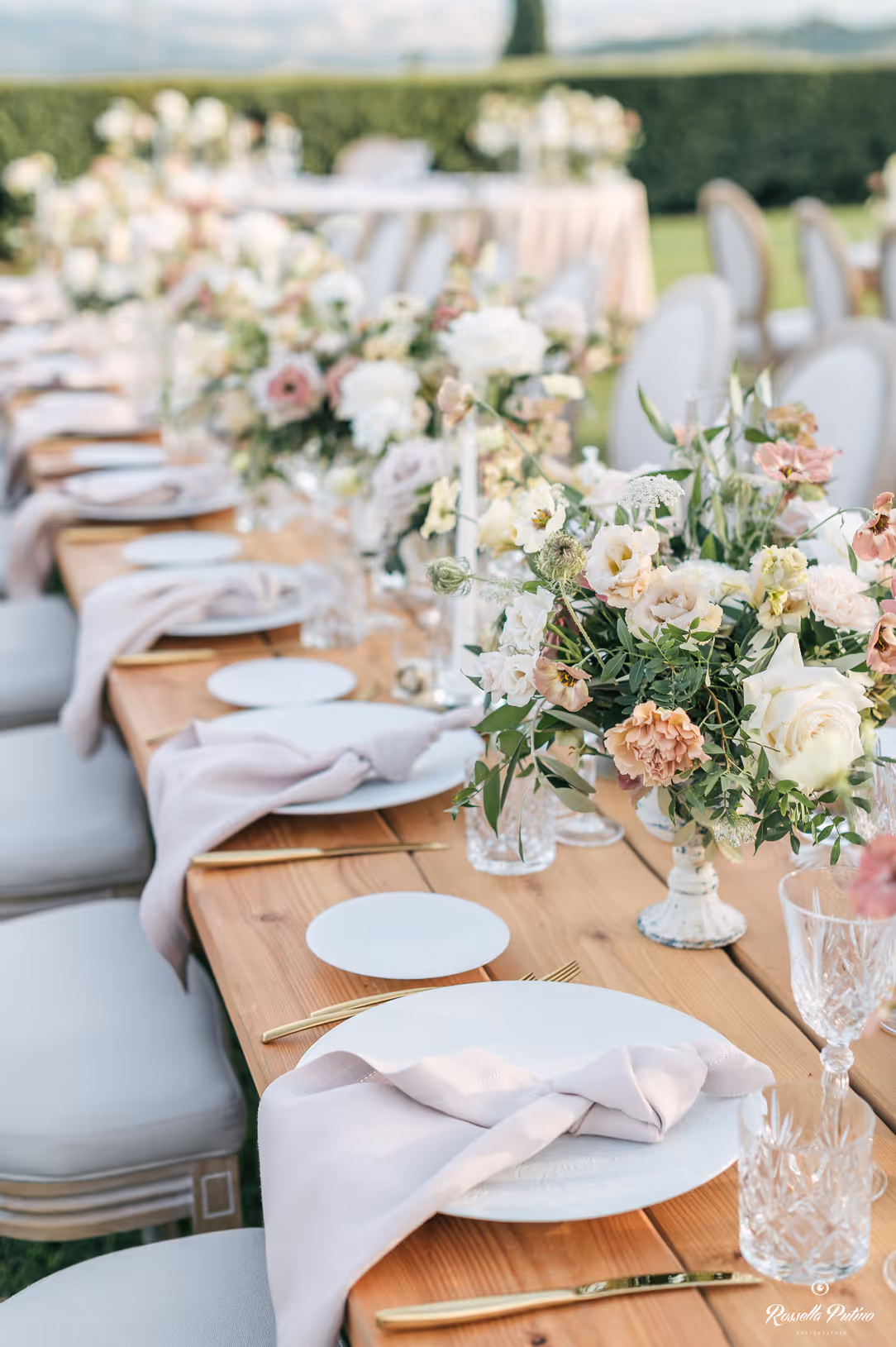 A close-up vertical photograph of a decorated wooden wedding table at Castello di Vicarello, showing knotted pale pink napkins on white plates, crystal glasses, and lush floral centerpieces with roses.