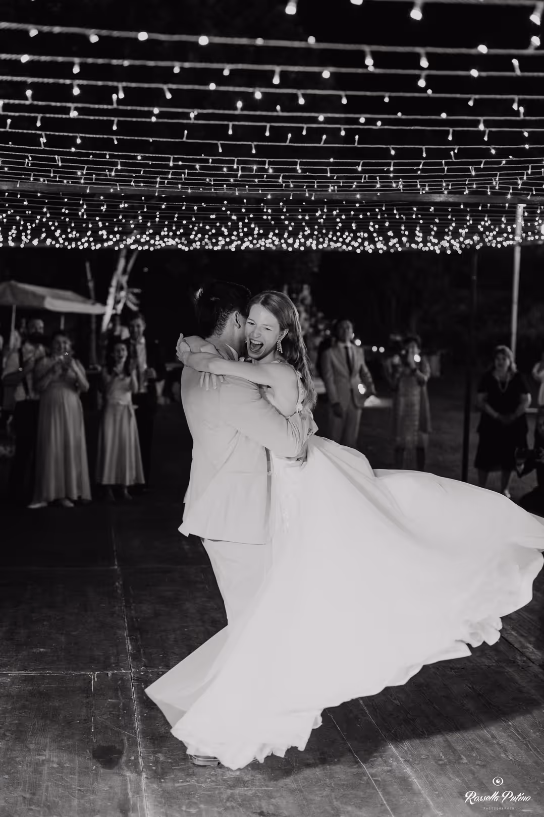 A dynamic vertical black and white wedding photograph of a groom spinning his laughing bride on an outdoor dance floor beneath a massive canopy of sparkling fairy lights.