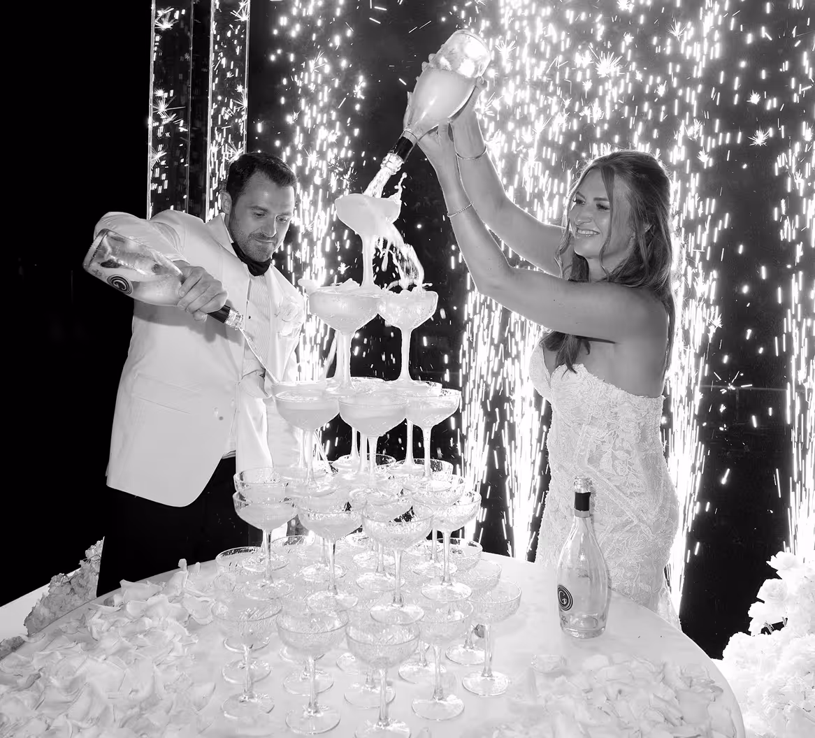 Foto in bianco e nero degli sposi mentre versano lo champagne su una monumentale piramide di calici di cristallo, con spettacolari fontane di fuochi d'artificio luminosi sullo sfondo durante la festa a Villa Erba.