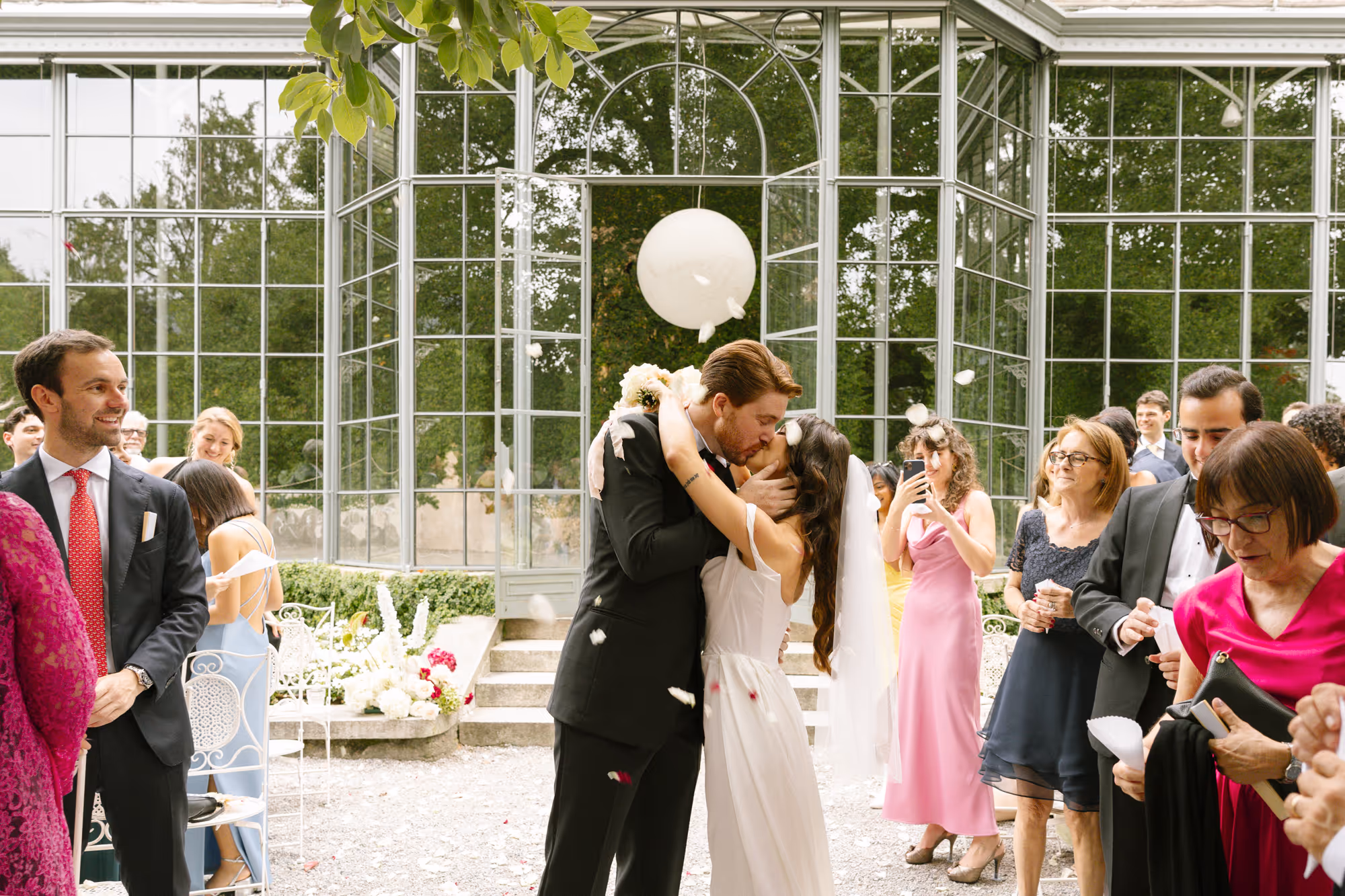 A bride and groom sharing a romantic kiss during their wedding recessional outside a historic glass greenhouse at Villa Grumello, with guests smiling and white petals falling around them.