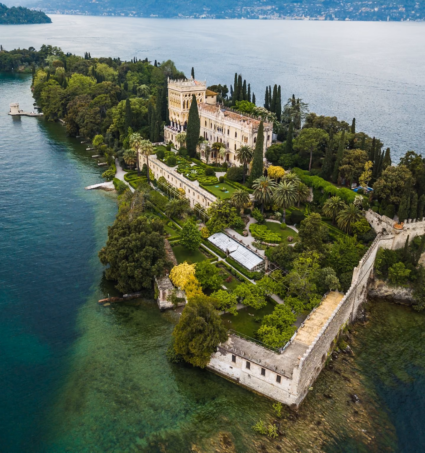 An aerial drone photograph of Isola del Garda on Lake Garda in Italy, featuring the historic Neo-Gothic villa, lush formal gardens, and ancient stone walls surrounded by clear turquoise water.