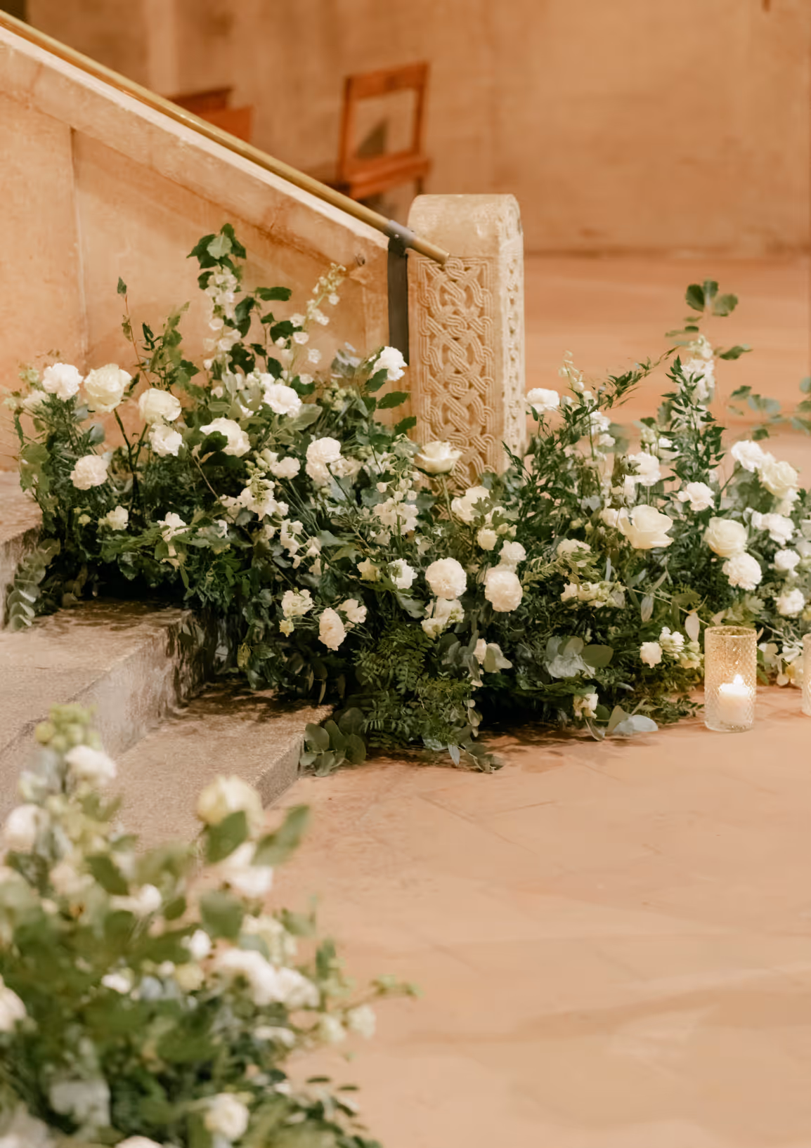 A vertical indoor photograph of lush white rose and green foliage floral installations lining the stone altar staircase inside a historic Italian church with a burning glass candle.