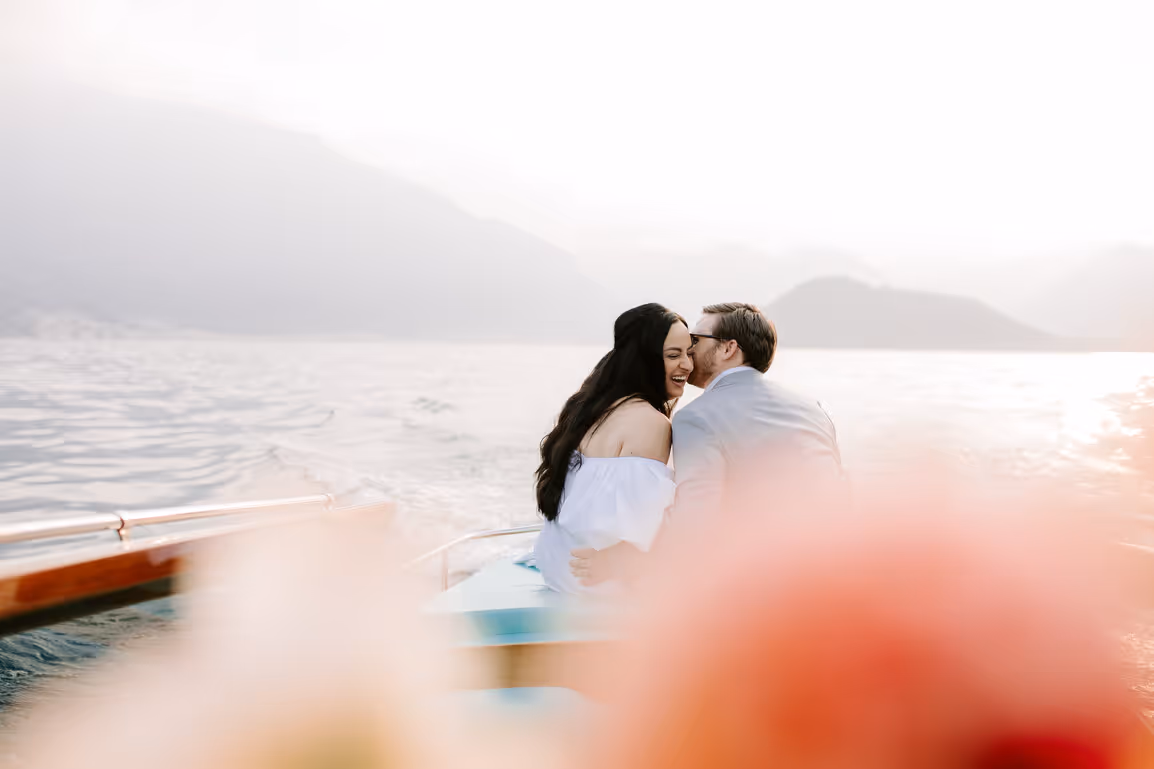 A sun-drenched horizontal wedding portrait of a bride and groom embracing on a luxury wooden boat on Lake Como with soft-focus peach flowers in the foreground.