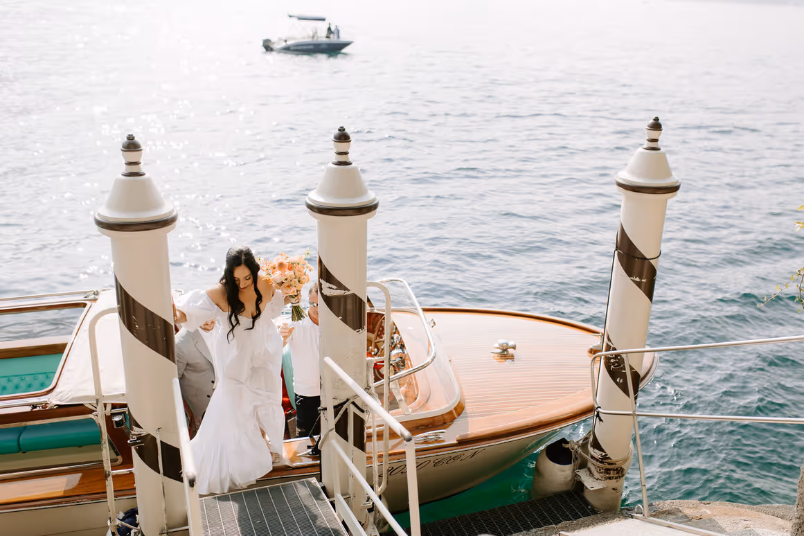 A high-angle photograph of a bride holding a peach bouquet as she steps off a classic wooden speedboat onto the stone dock of Villa del Balbianello on Lake Como.