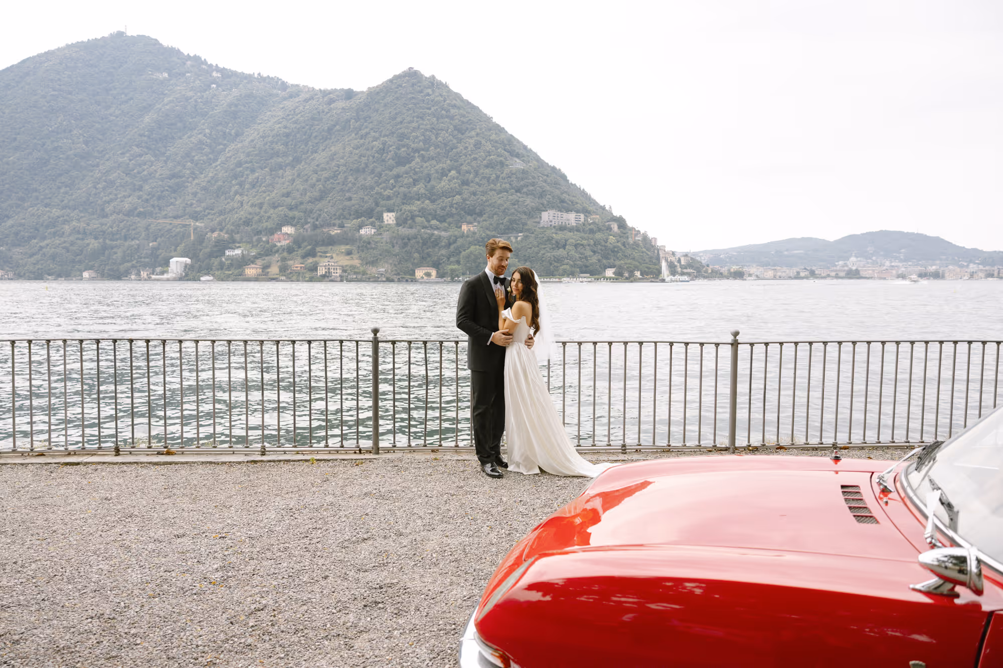A wide-angle portrait of a bride and groom by the lakeside railing at Villa Grumello, with a scenic background of Lake Como and mountains, and the hood of a red vintage car in the foreground.