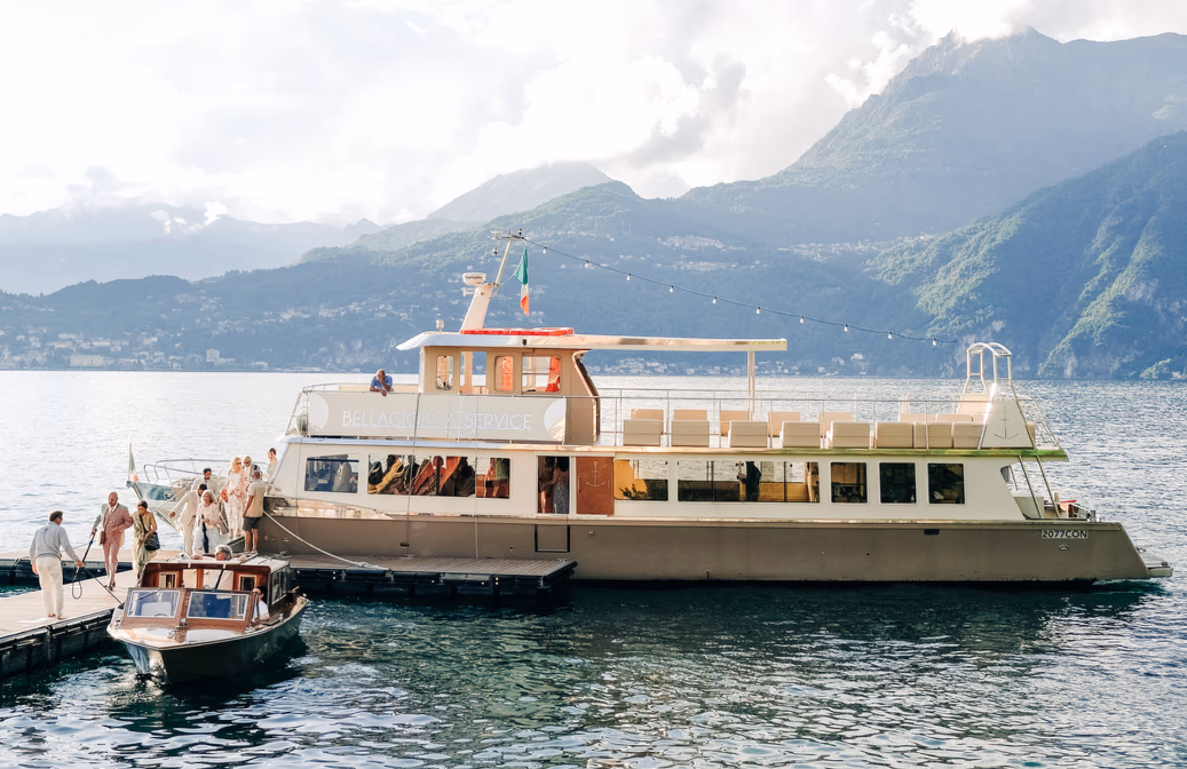 A wide landscape photograph of wedding guests boarding a luxury private cruise boat from Bellagio Boat Service at a pier on Lake Como, with a classic wooden speedboat nearby and sunlit mountains in the background.