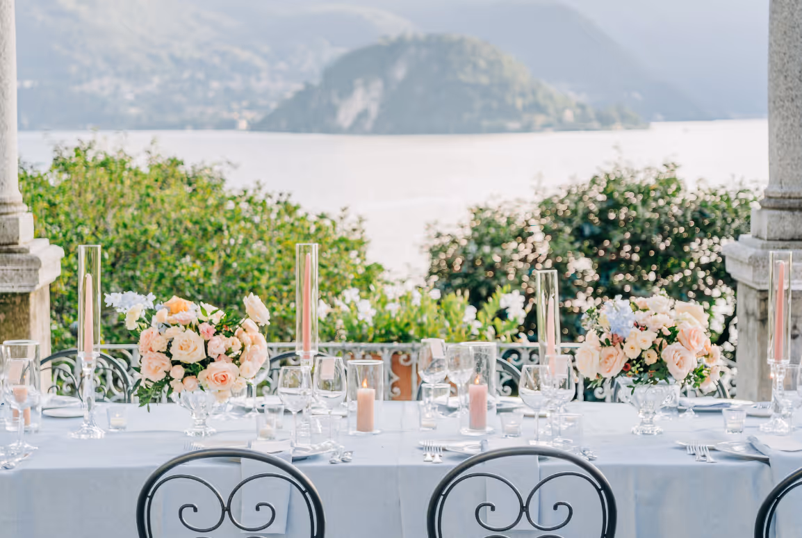 A scenic landscape view of a wedding reception terrace overlooking Lake Como and distant mountains, framed by elegant dining tables with pink candles and floral arrangements.