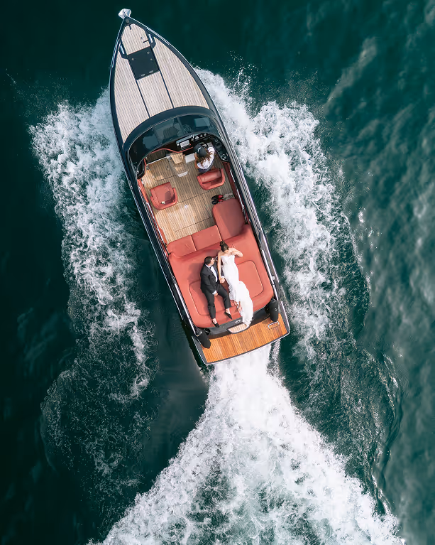 Aerial drone view of a bride and groom relaxing on a luxury wooden speed boat cruising on the deep blue water of Lake Como.