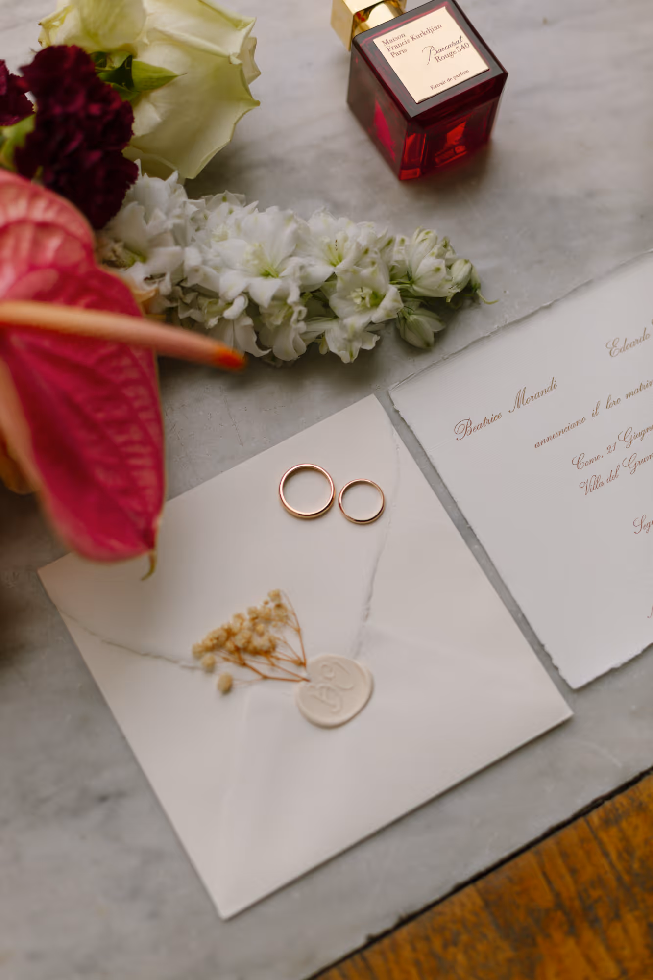 A luxury wedding flat lay featuring custom deckled-edge invitations, gold wedding bands on a white envelope with a wax seal, a bottle of Baccarat Rouge 540 perfume, and fresh floral accents.