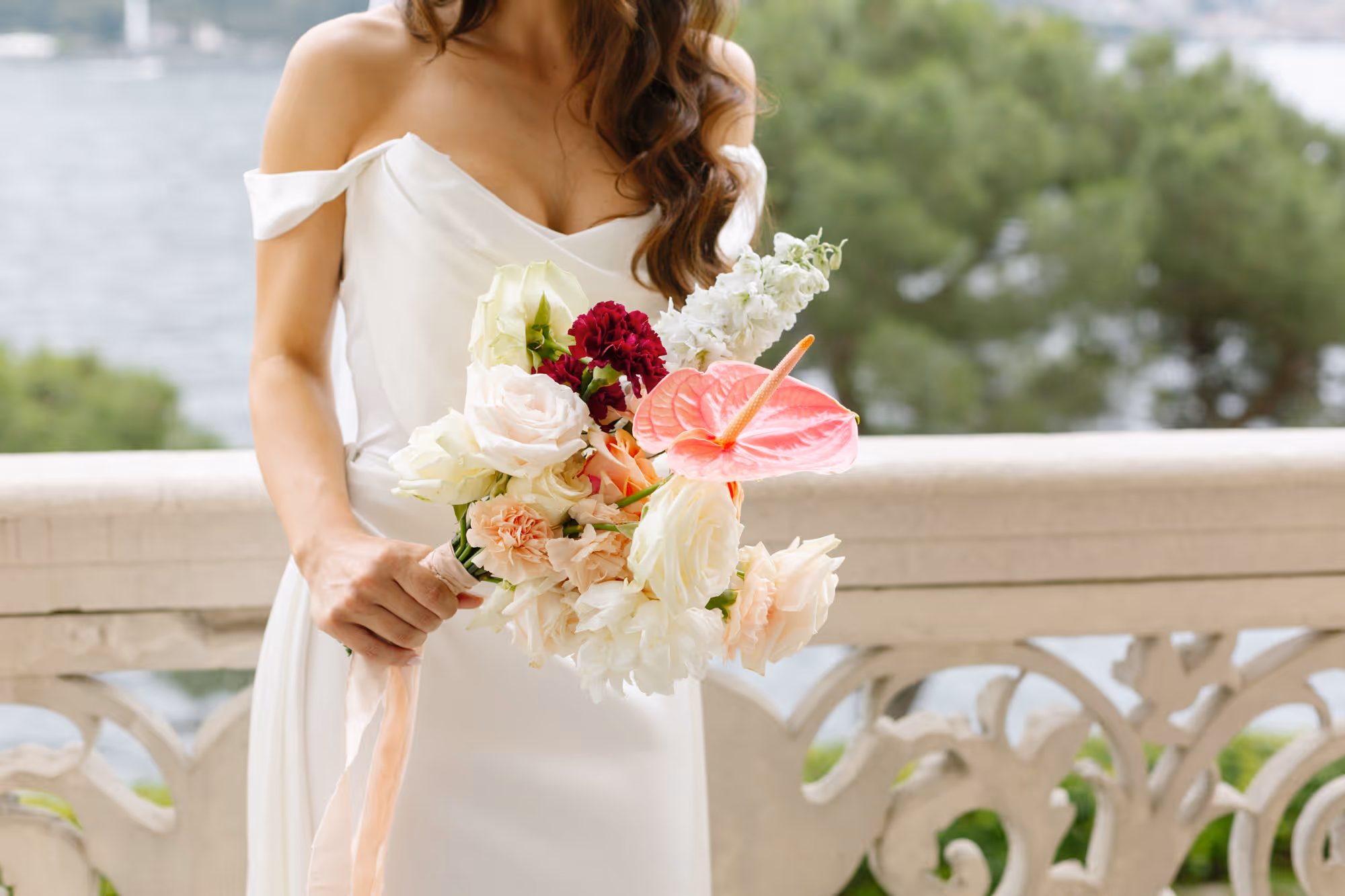 A close-up shot of a bride in an elegant off-the-shoulder gown holding a modern wedding bouquet with white roses, peach carnations, and a striking pink anthurium flower, standing by a stone balcony at Villa Grumello.