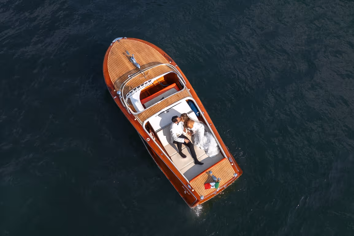 Veduta aerea dall'alto di un motoscafo in legno che naviga sul Lago di Como vicino a Villa Erba, con gli sposi eleganti distesi a poppa mentre si scambiano un bacio.
