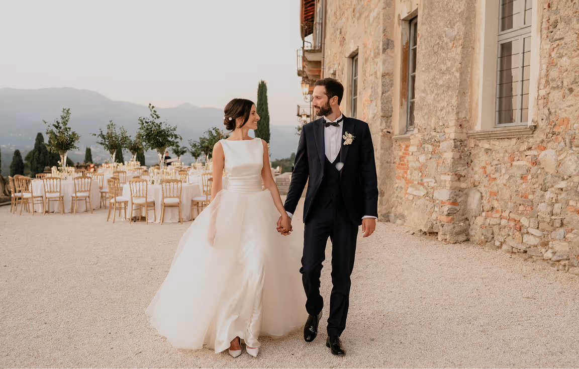 Sposi eleganti che camminano mano nella mano accanto alle mura in pietra di una dimora storica, con tavoli tondi allestiti per il ricevimento sullo sfondo al Castello Durini.