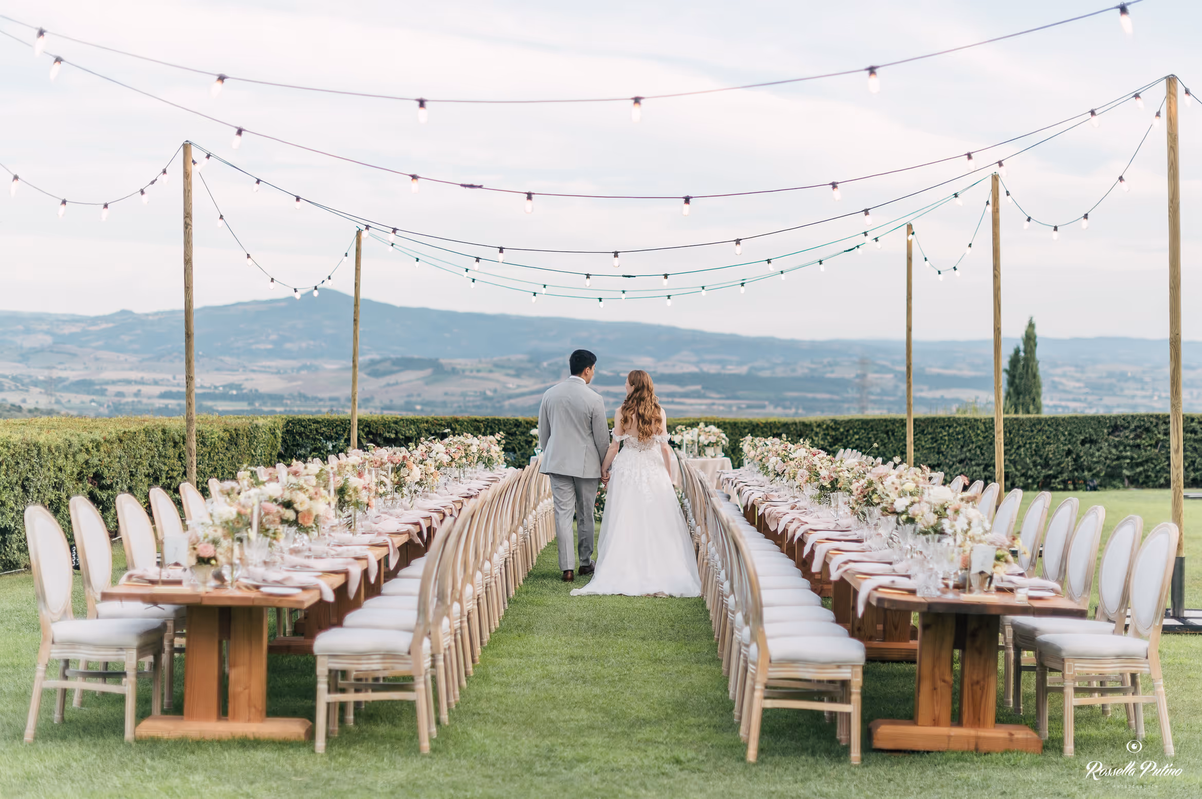 A wide-angle photograph of an outdoor wedding reception at Castello di Vicarello, featuring two long wooden banquet tables set on a green lawn under strings of bistro lights, with the bride and groom walking down the center aisle looking out at the Tuscan hills.