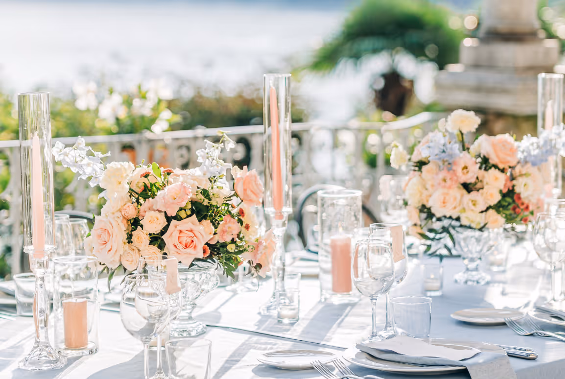A bright outdoor wedding reception table setting featuring lush peach and pink floral centerpieces and tall glass candle holders with pastel taper candles on a lakeside terrace.