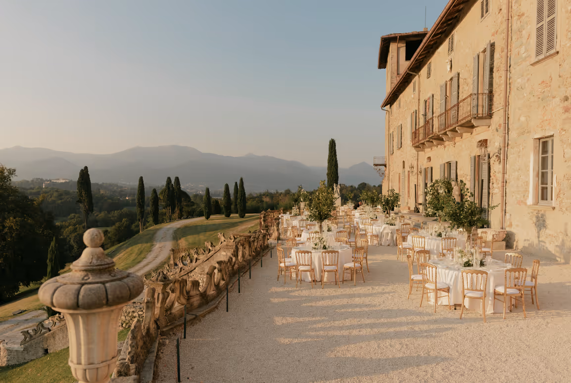 An expansive outdoor wedding reception setup at Castello Durini with round dining tables and gold chairs on a scenic gravel terrace overlooking the Italian countryside hills and mountains.